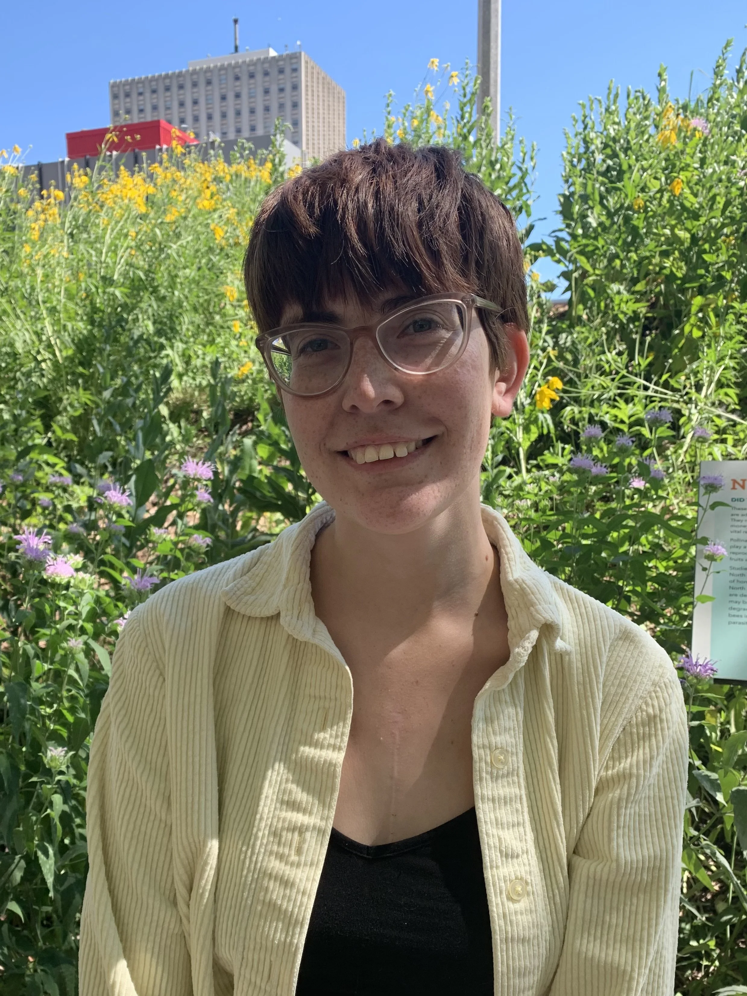 A woman with short auburn hair, wearing glasses, a black top, and a light yellow corduroy shirt, outdoors with green plants, a city building, and blue sky in the background.