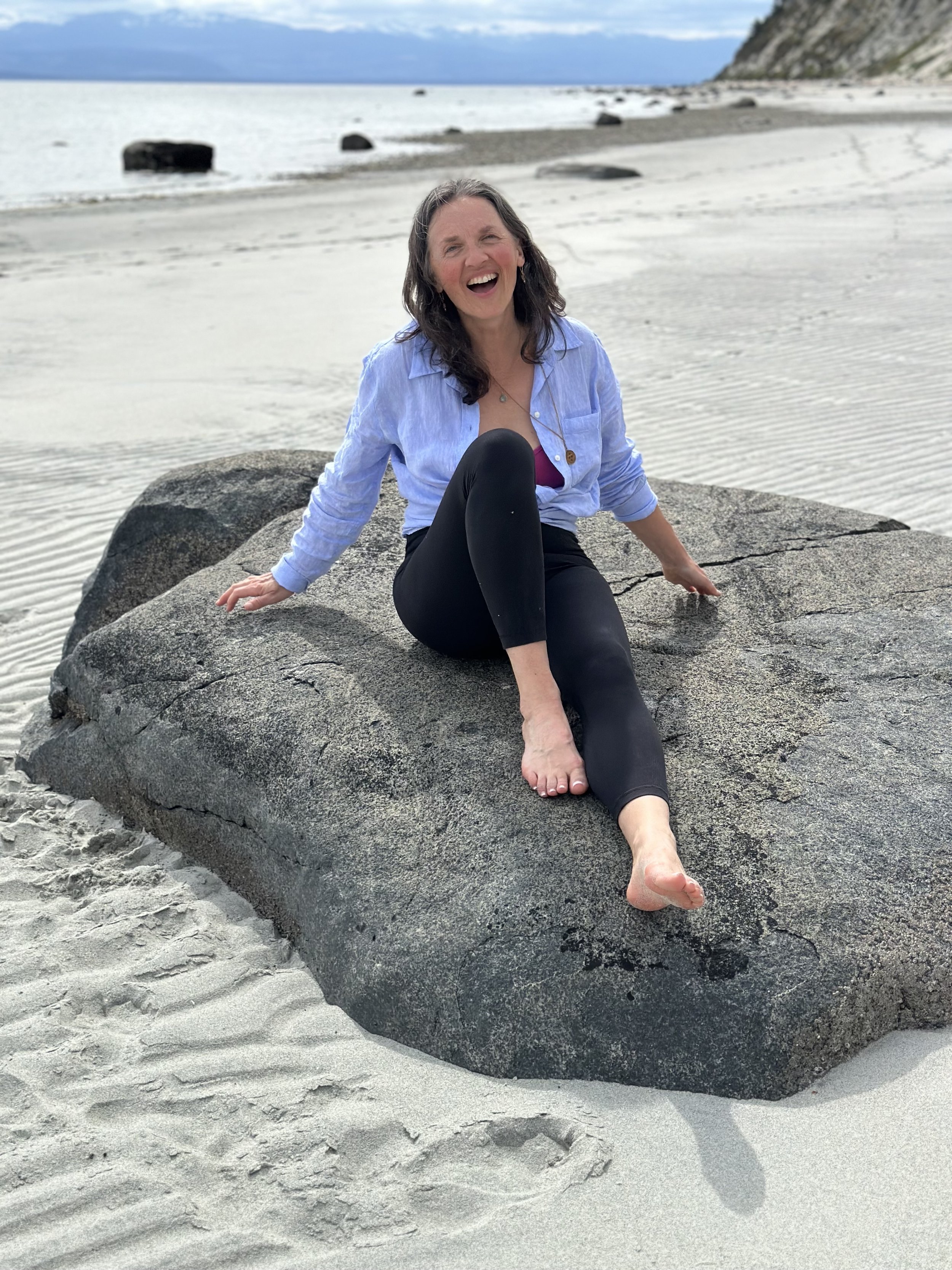 A woman sitting on a large rock at the beach, smiling and laughing, with a sandy shoreline and ocean in the background.