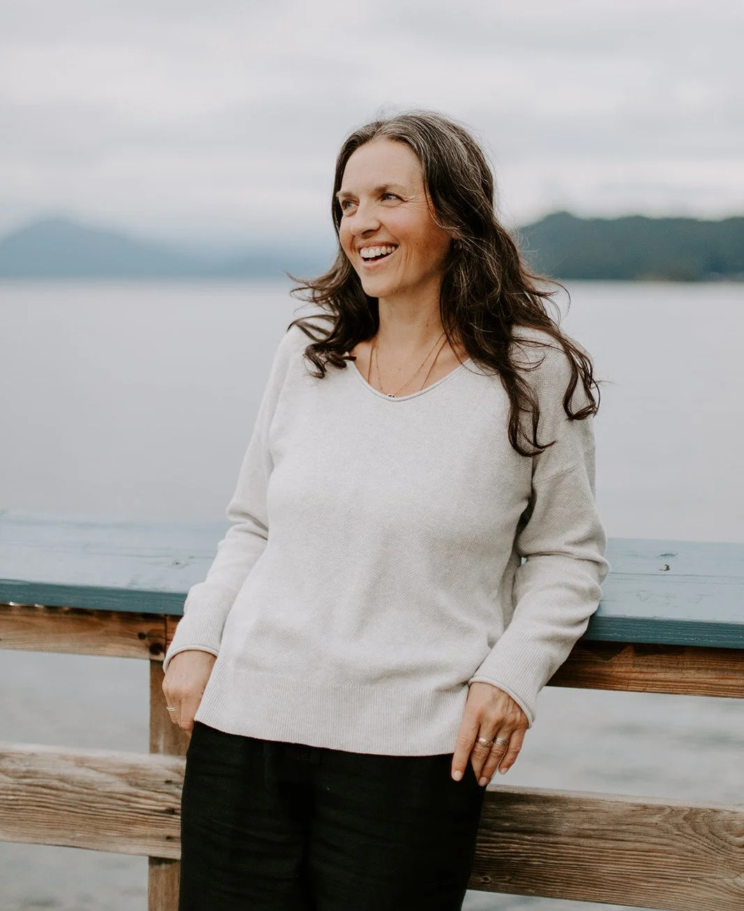 A woman standing outdoors by a lake, smiling and looking to the side, with mountains and cloudy sky in the background.