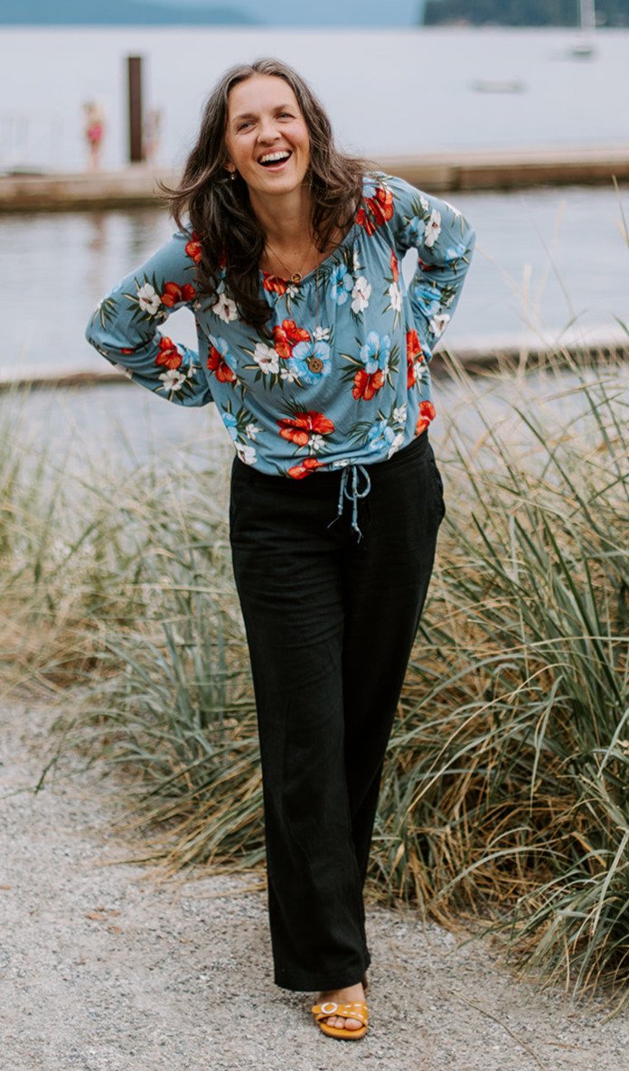 A woman in a floral blue top and black pants, smiling and standing outdoors near a body of water with grasses and a dock.