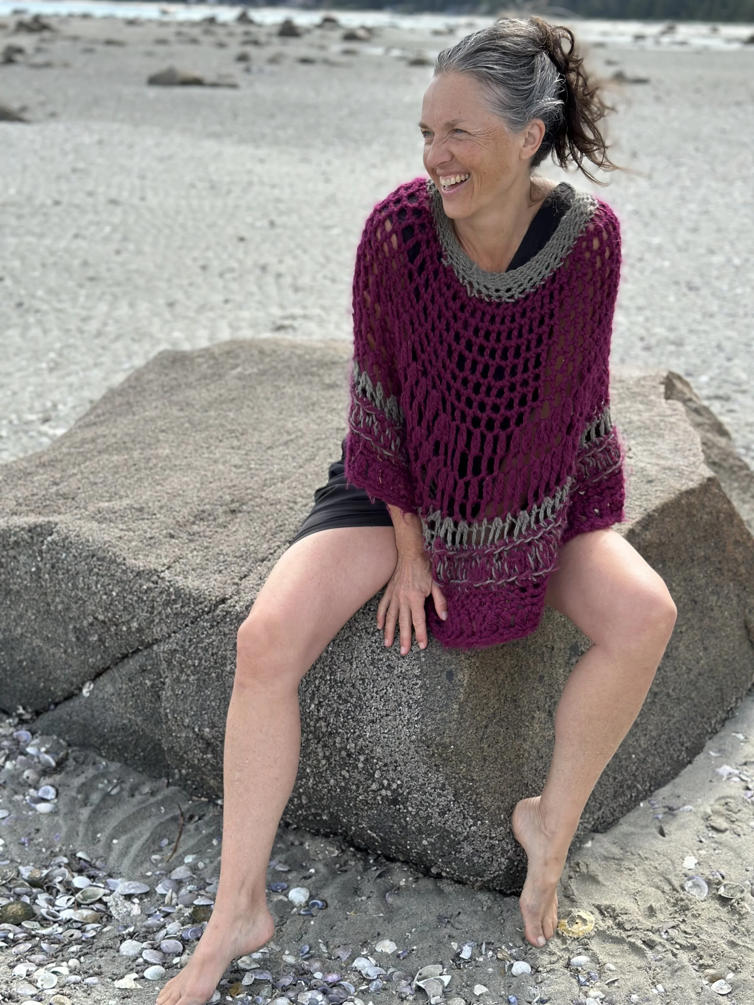 A woman with grey hair in a ponytail, wearing a pink and grey crochet sweater, is sitting on a large rock on a beach with shells on the sand, smiling and looking to the side.