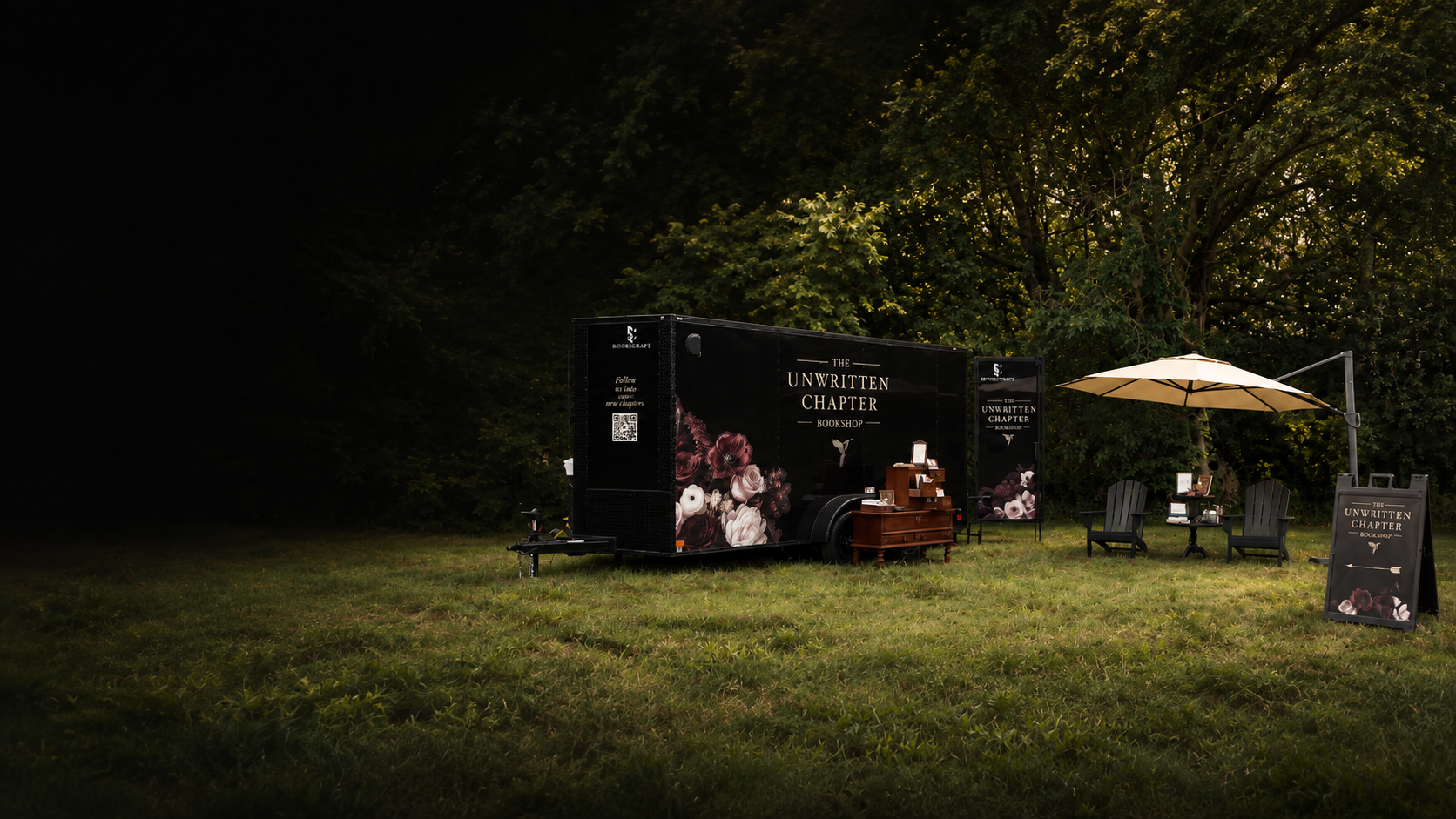 A mobile bookshop called 'The Unwritten Chapter' set up outdoors on a grassy area with seating, an umbrella, and a sign, with trees in the background.