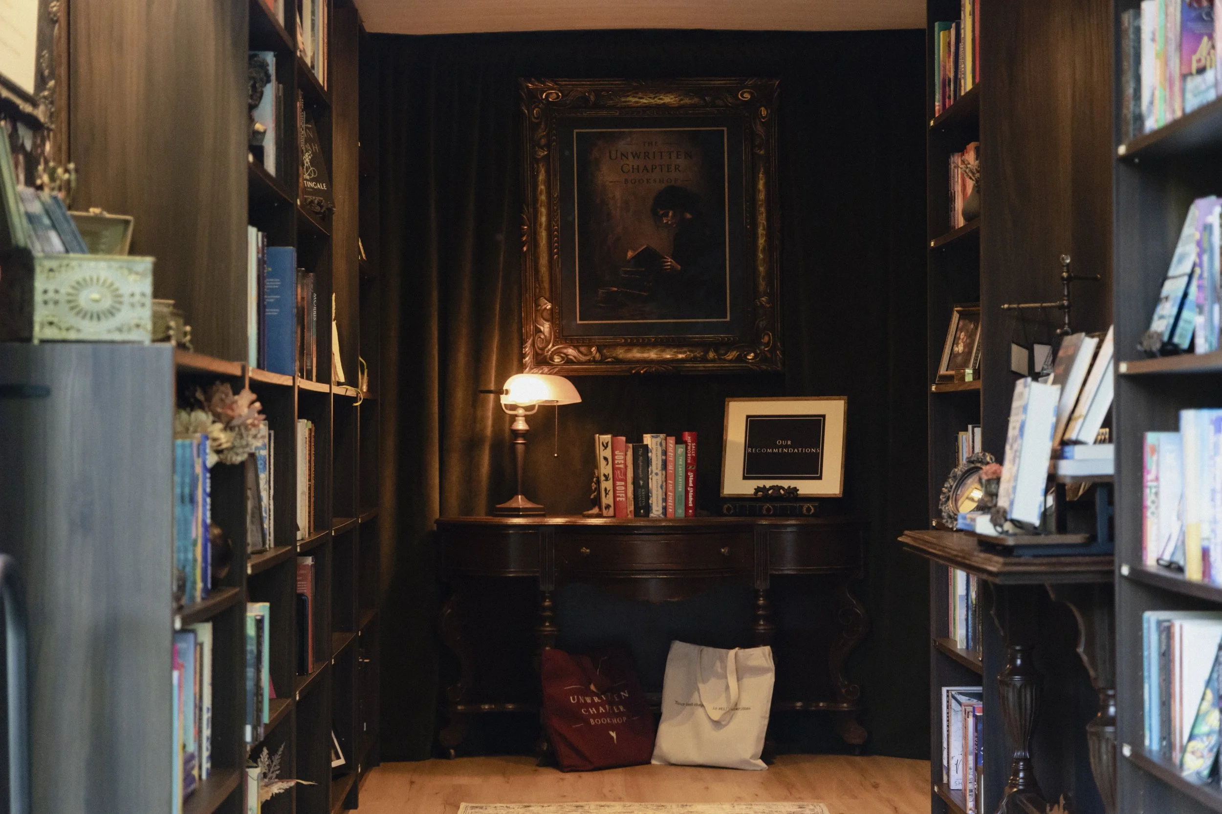 A cozy bookstore with bookshelves on both sides, a small table in the back with bags on the floor, a framed poster titled 'The Unwritten Chapter', a small lamp, and books on display.