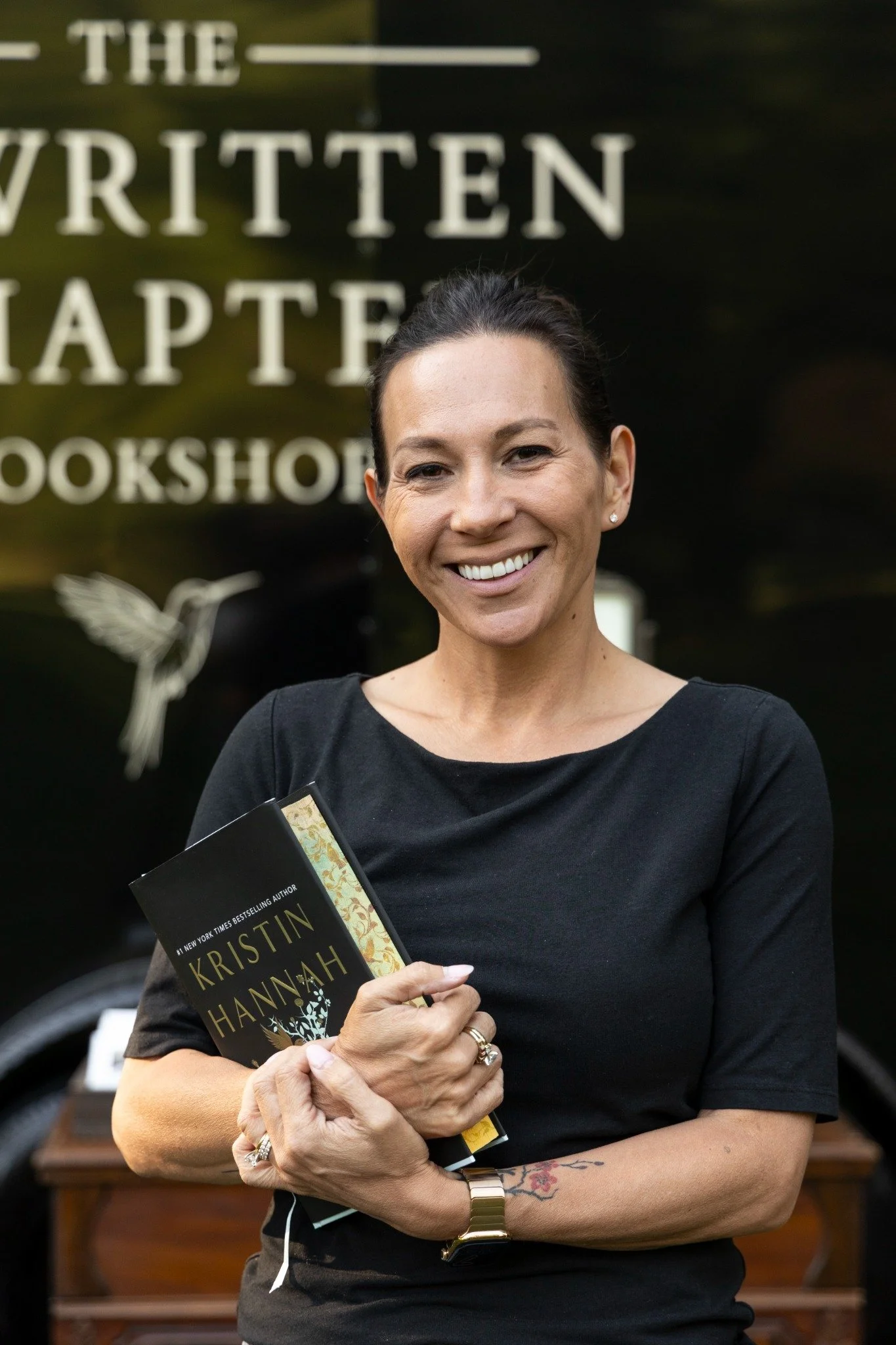 A smiling woman with dark hair pulled back, wearing a black top, holding a book titled 'The Seven Husbands of Evelyn Hugo' by Kristin Hannah, standing outdoors in front of a dark sign with white text and a small white bird illustration.