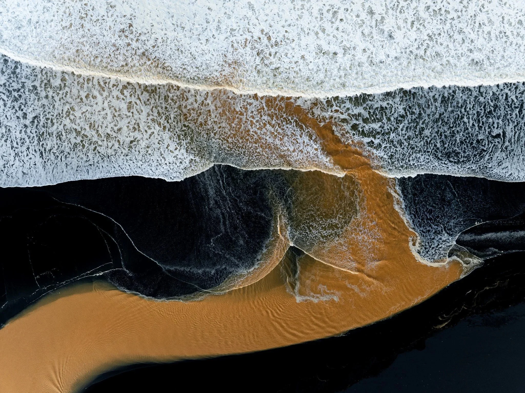 Aerial view of a brown glacial river meeting the ocean across a black sand beach in Iceland