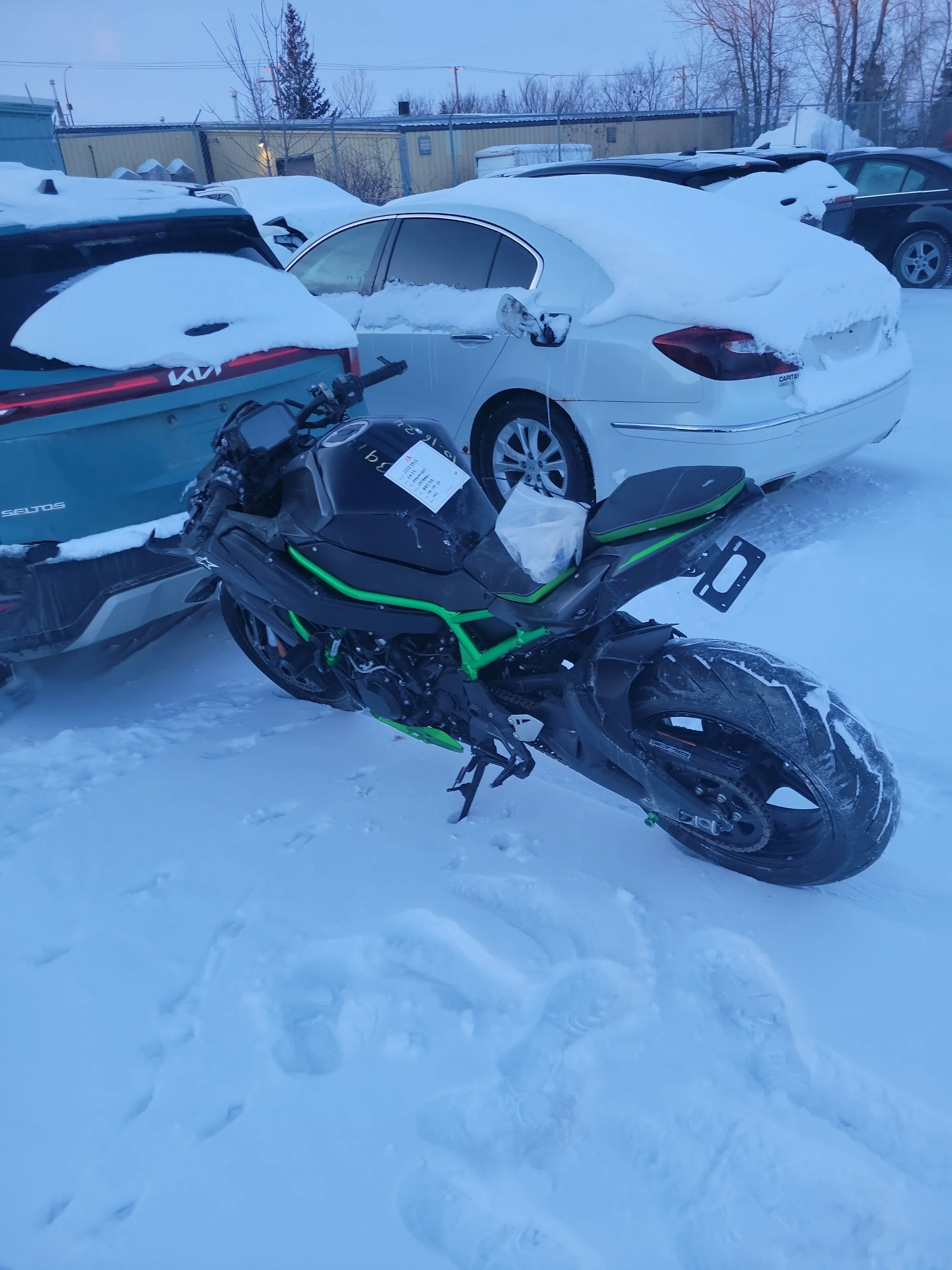 A snow-covered motorcycle and cars parked in a snow-covered lot. The motorcycle is black and green, and the cars are partially covered in snow.