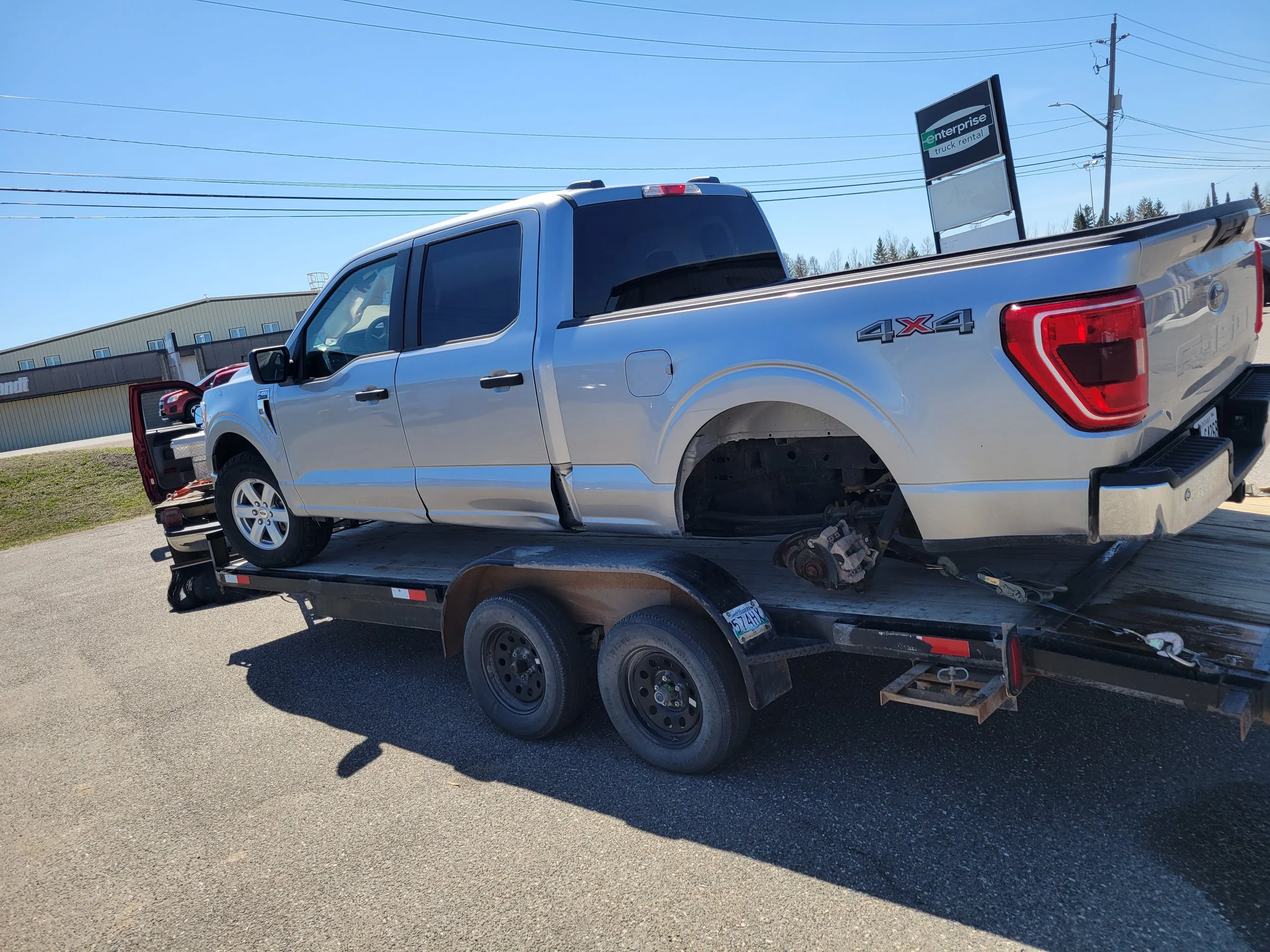 A silver Ford F-150 pickup truck with 4x4 marking on the side, missing wheels, loaded onto a flatbed tow truck with its side door open, parked outside near a commercial building with an 'Enterprise' truck rental sign and a row of building windows in 