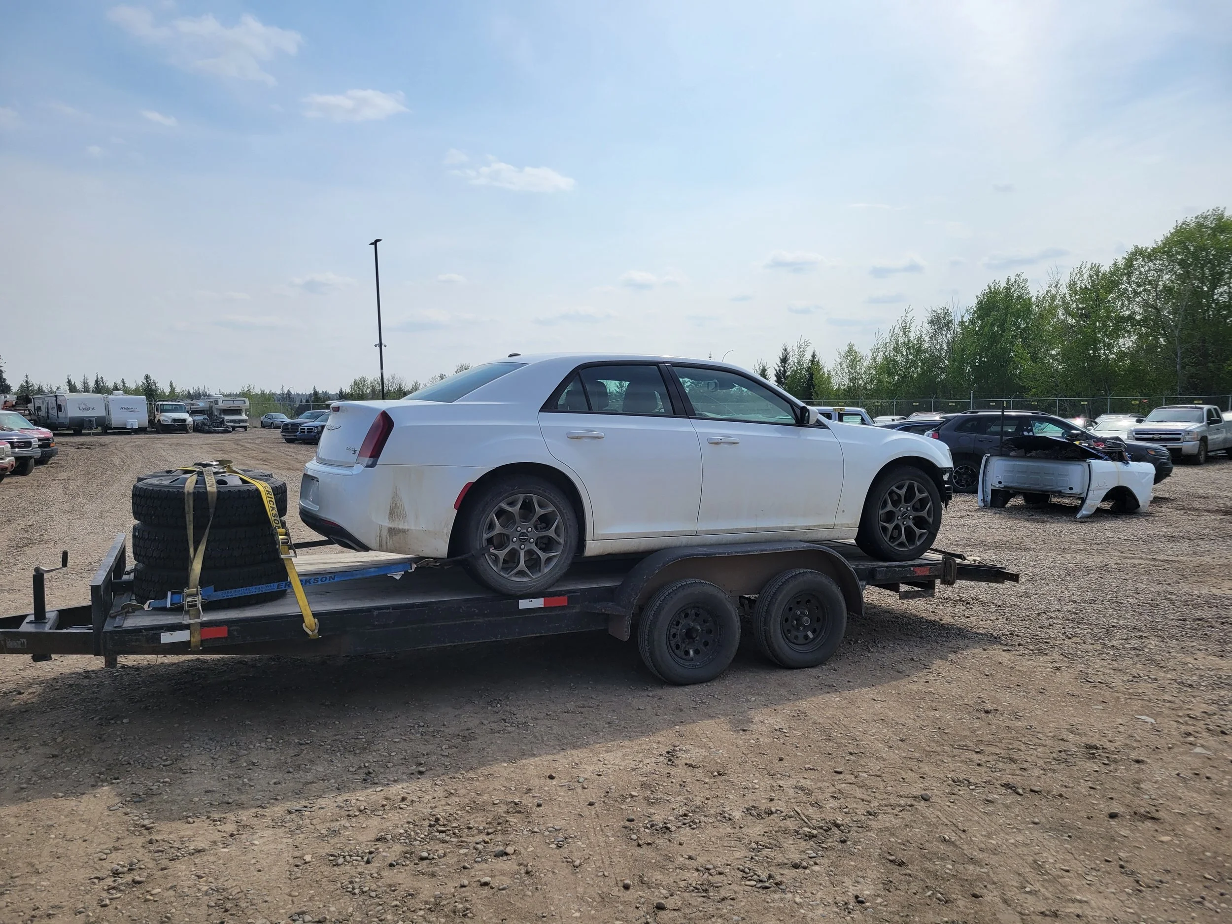 A white sedan car loaded on a trailer with tires and equipment, parked in a gravel lot near other vehicles and surrounded by trees under a partly cloudy sky.