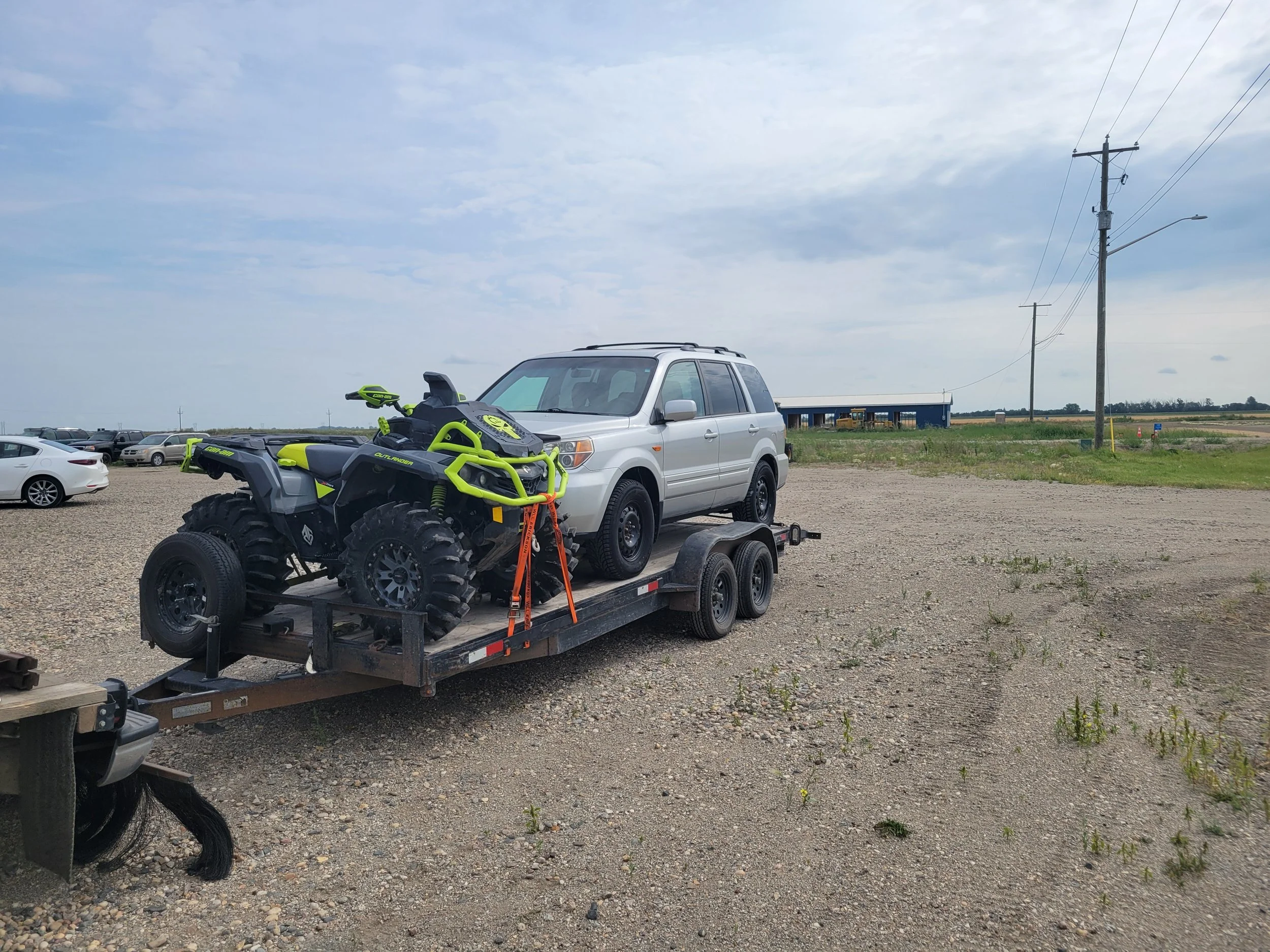 A black ATV on a trailer hitched to a silver SUV in a gravel parking lot, with several parked cars and power lines under a cloudy sky.