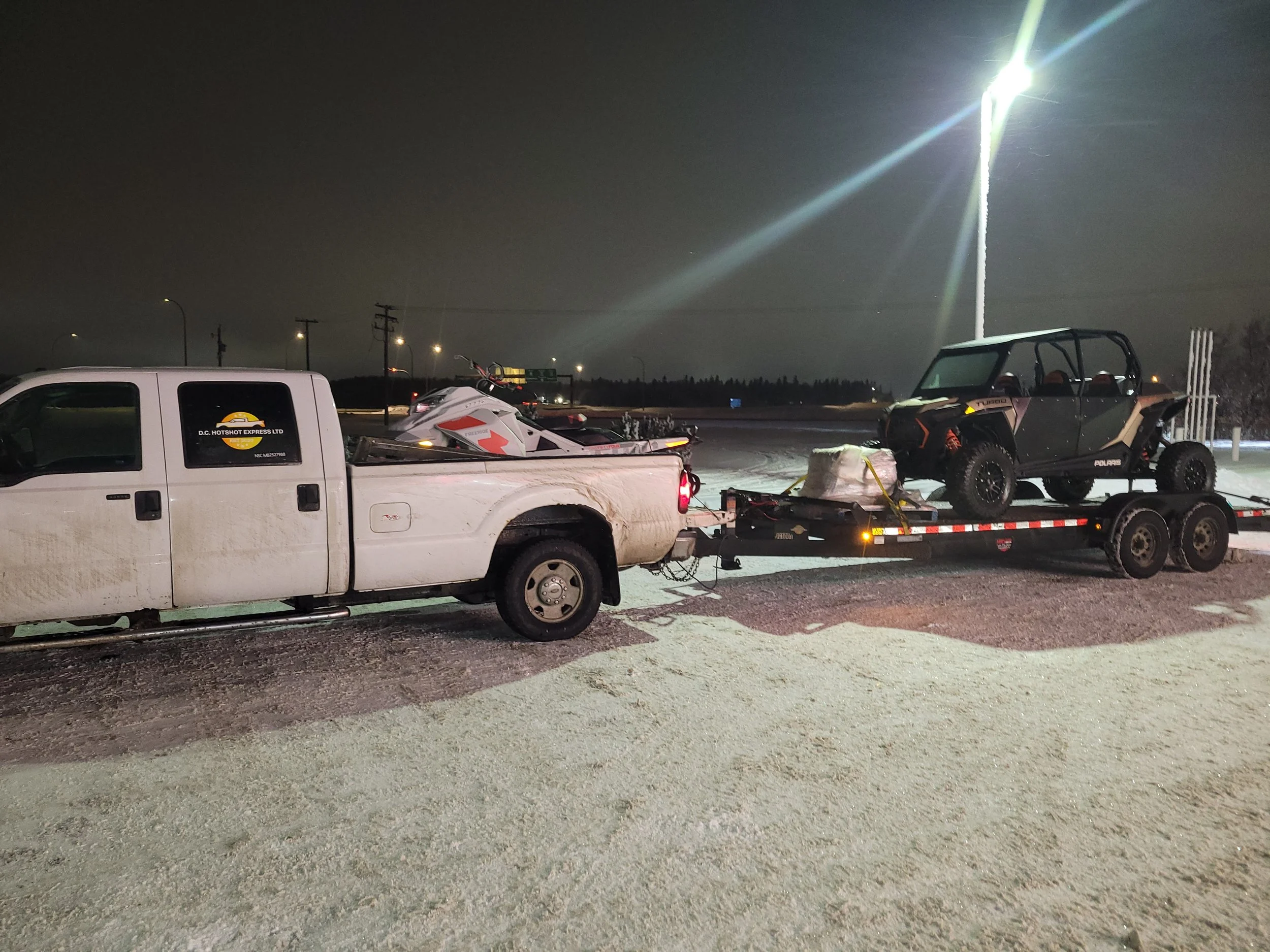 Night scene at a parking lot with vehicles, including a white pickup truck carrying a Polaris off-road vehicle and a jet ski, with street lights and a bright light source in the sky.
