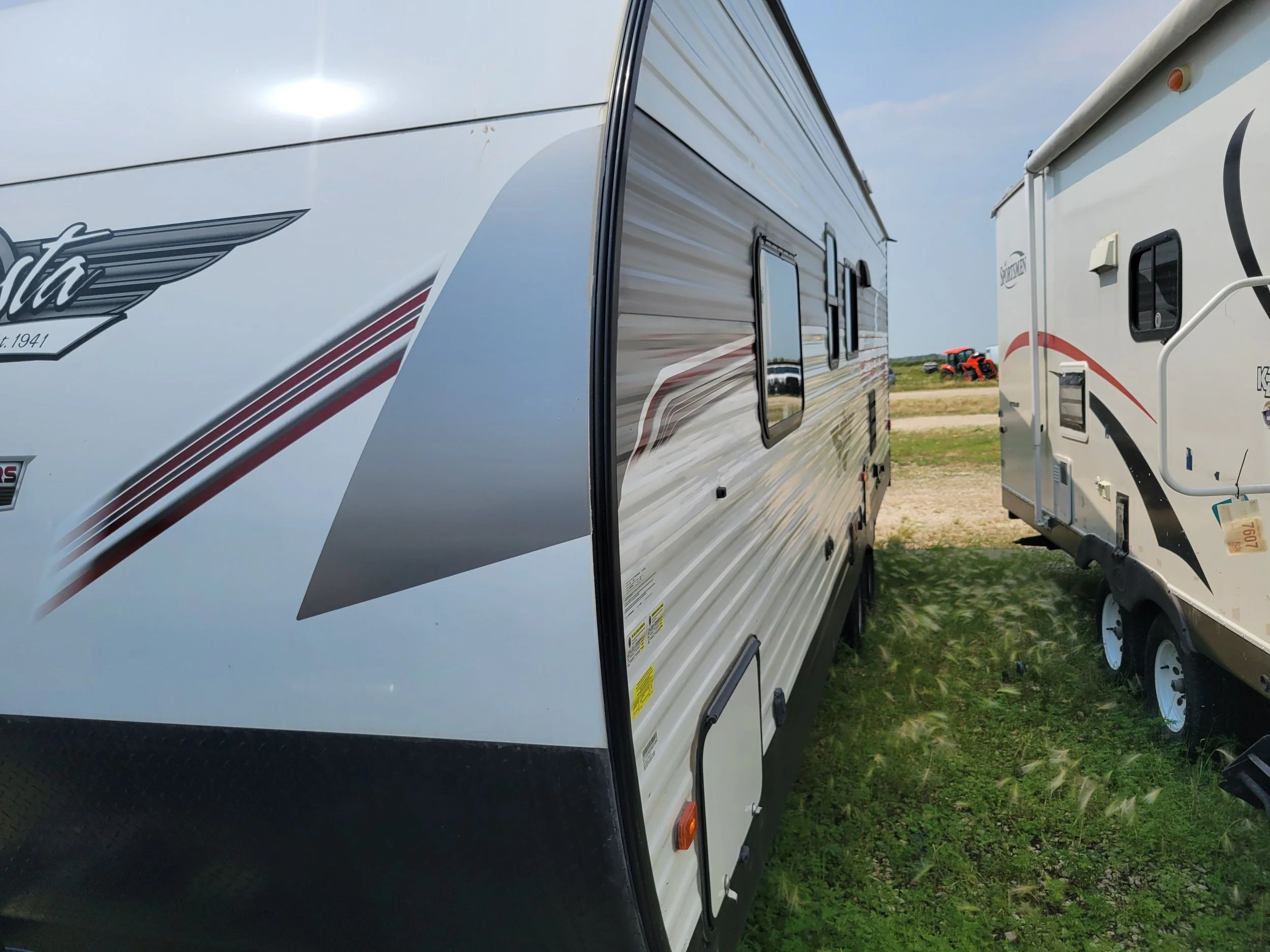 Two travel trailers parked on grass with a cloudy sky, and a red tractor in the background.