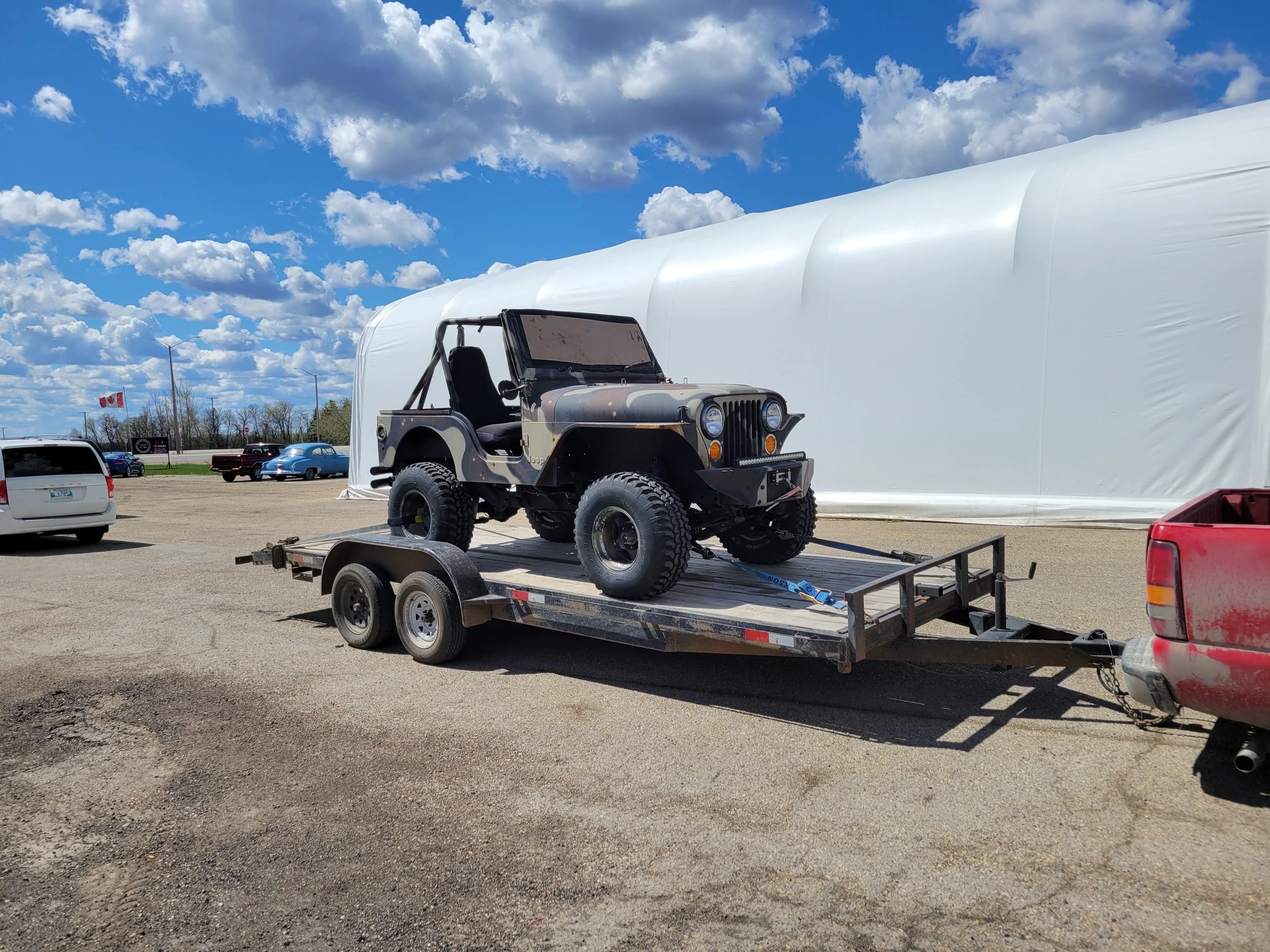 A camouflage Jeep on a flatbed trailer in a parking lot with multiple cars and a large white tent structure in the background. Blue sky with scattered clouds and streetlights visible.