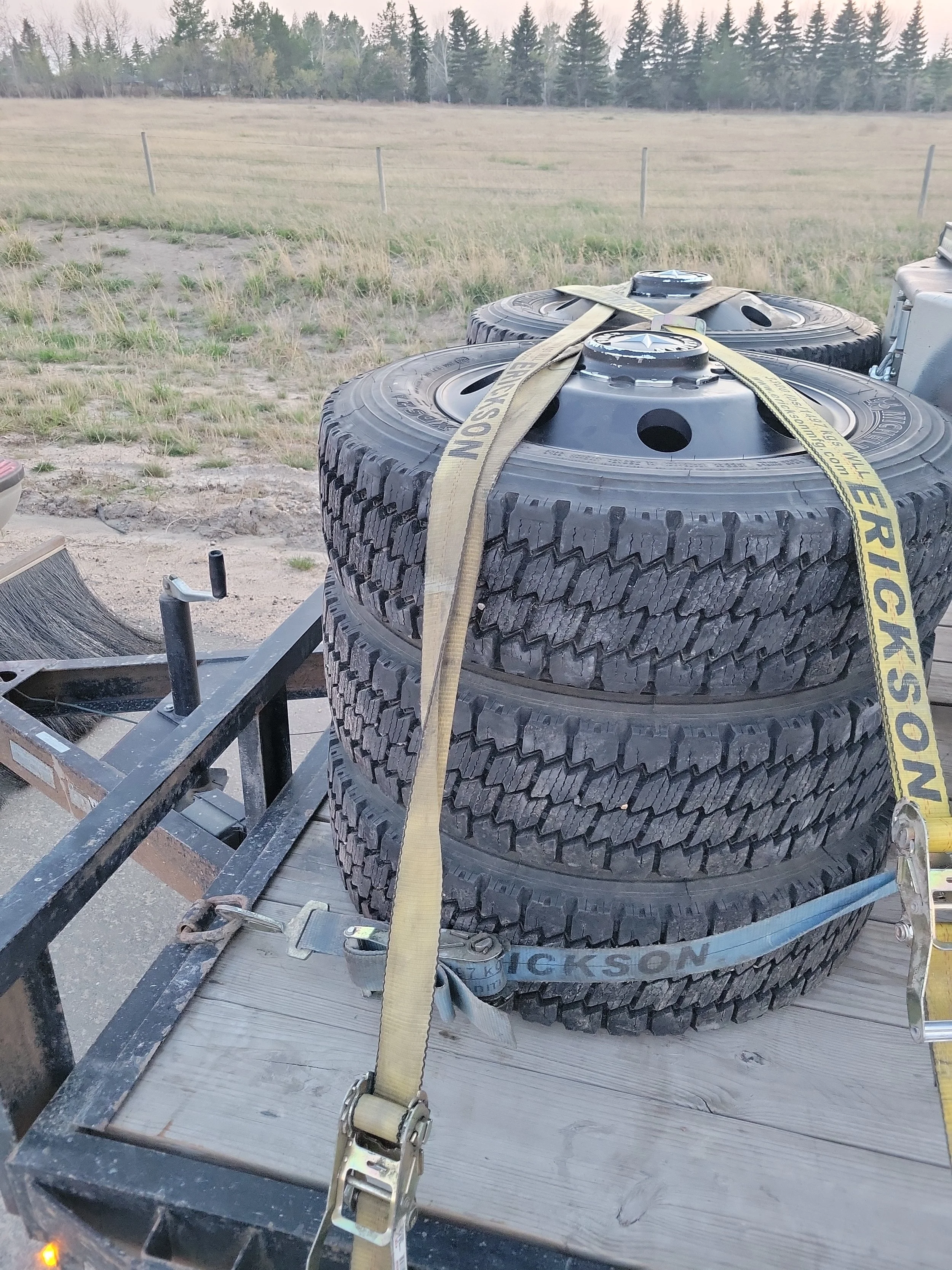 Stacks of four car tires secured with yellow ratchet straps on a flatbed trailer, in an outdoor field.