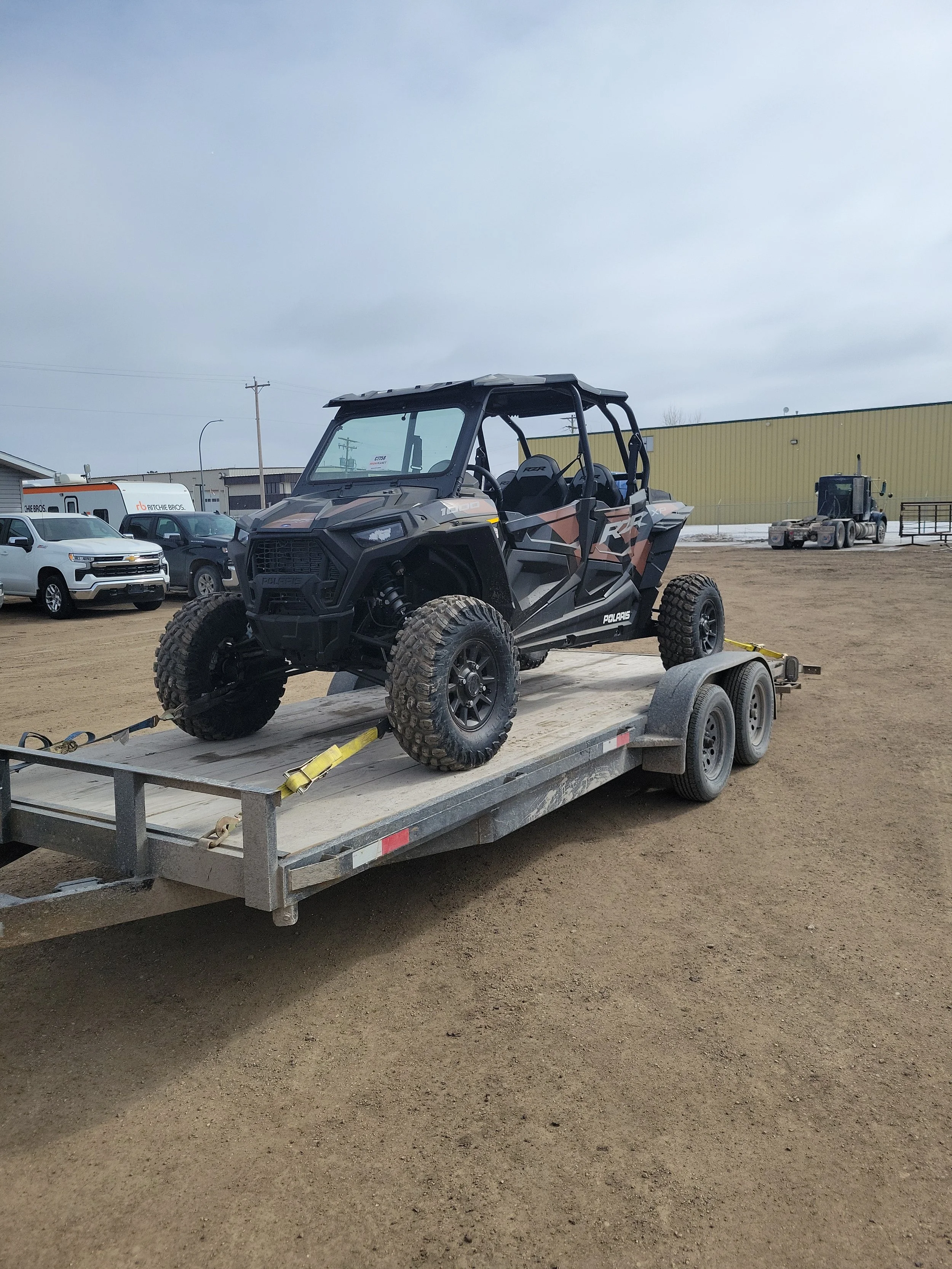 A Polaris RZR off-road vehicle on a flatbed trailer parked on dirt at an outdoor lot with other vehicles and a yellow industrial building in the background.