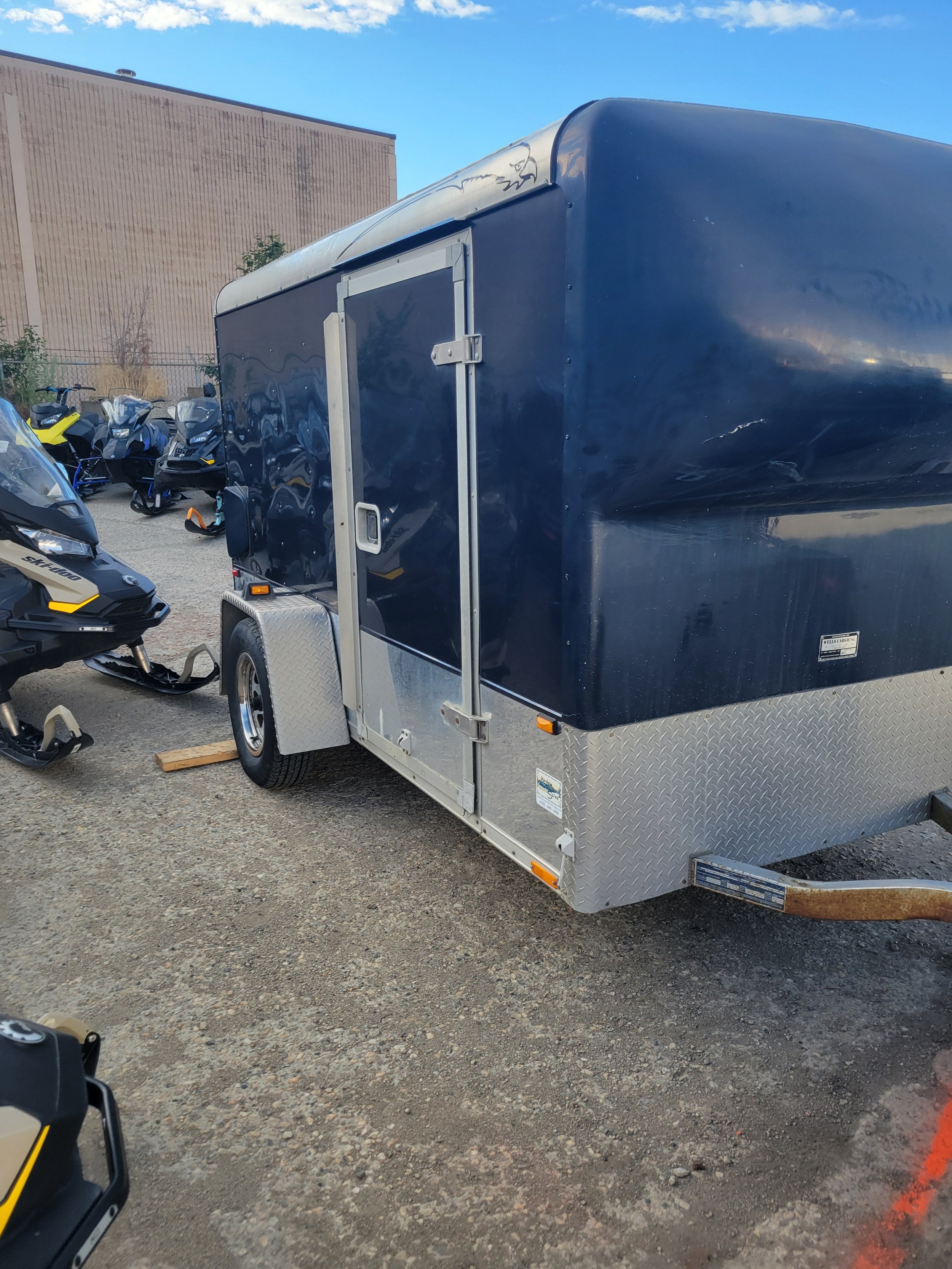 Black enclosed trailer with a silver front, parked on gravel. There are snowmobiles and ski-doos nearby, and a building in the background under a clear blue sky.