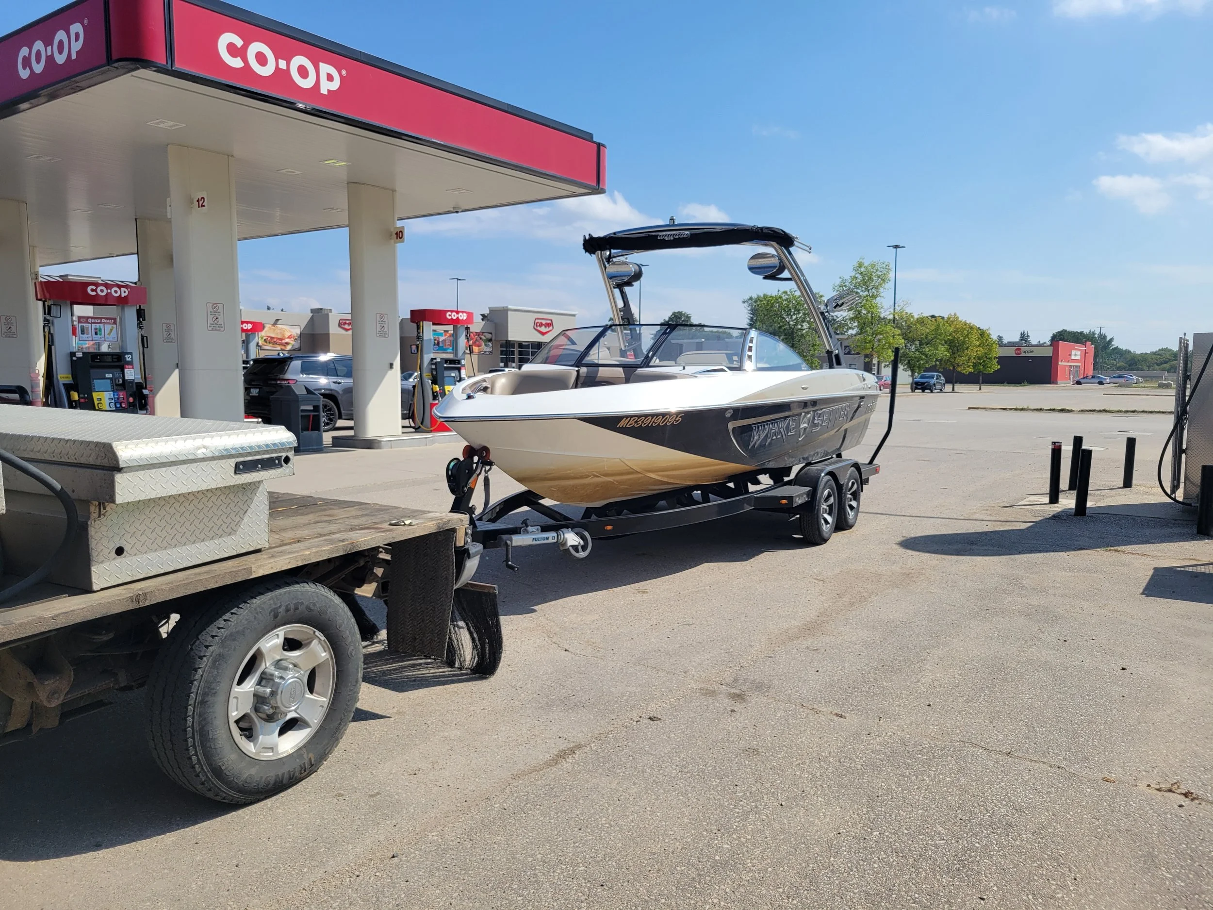 A boat on a trailer at a Co-op gas station under a blue sky with scattered clouds. The gas station has multiple fuel pumps and a canopy with the Co-op logo. There are cars and a building with a red and gray exterior in the background.