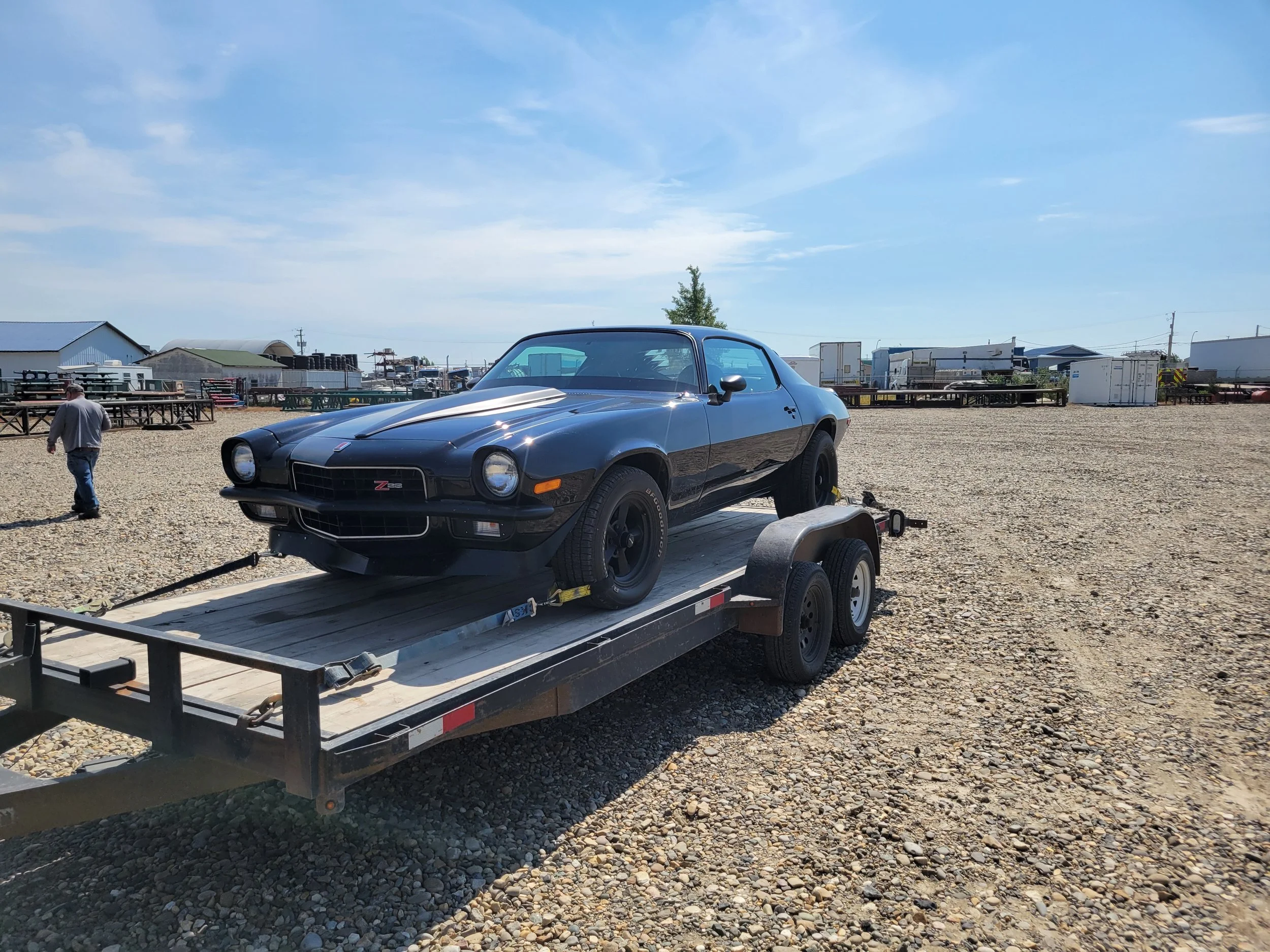 A classic black Chevrolet Camaro Z28 car on a trailer in an outdoor lot with gravel ground, surrounded by various industrial buildings and equipment, under a partly cloudy sky.