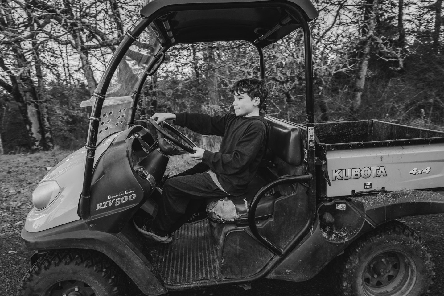 A young boy sitting in the driver's seat of a utility vehicle in a wooded area.