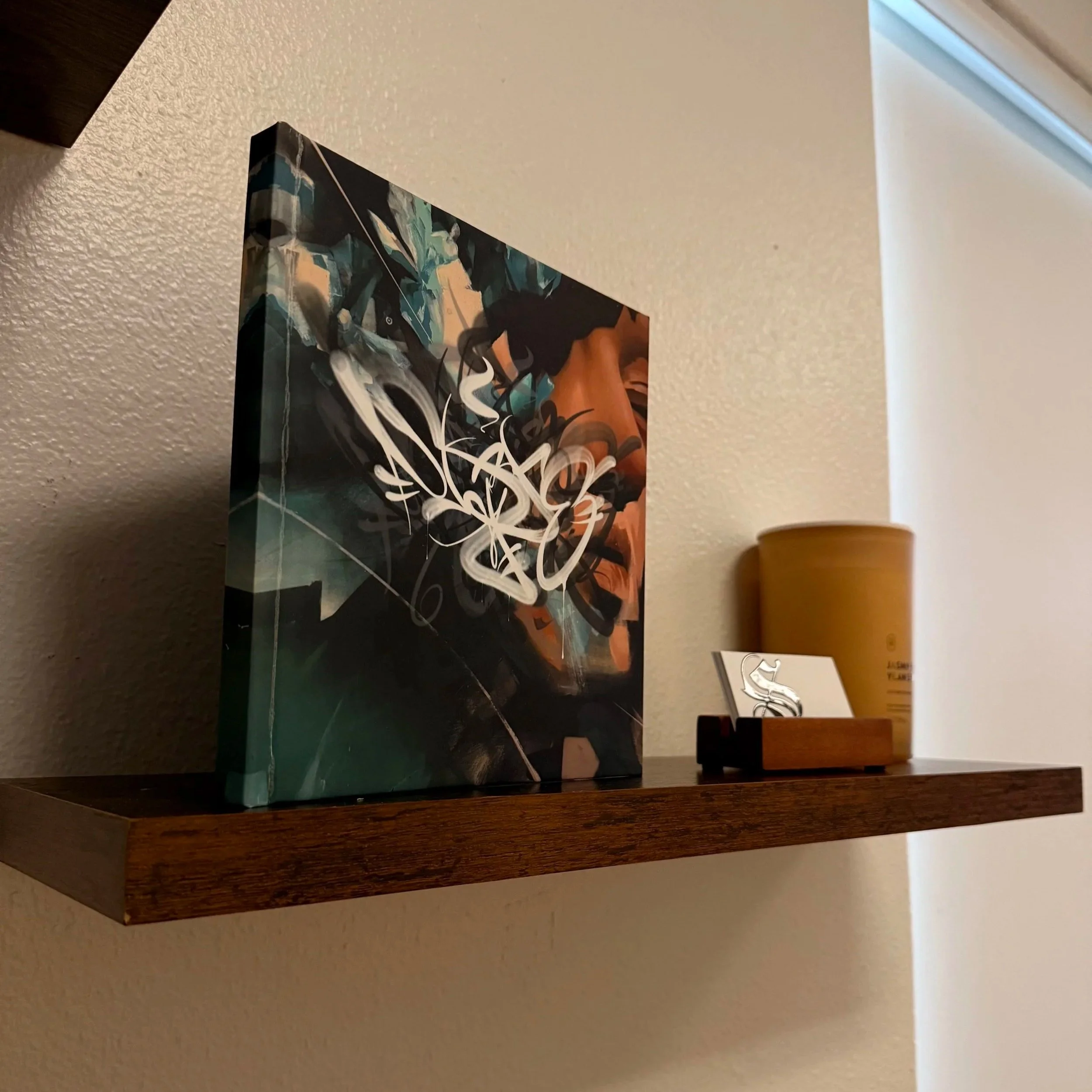 A colorful abstract painting on a small wall hanging or canvas placed on a shelf or ledge against a beige textured wall. To the right, there is a yellow object, possibly a container or a cup. Part of a white object or label is also visible nearby.