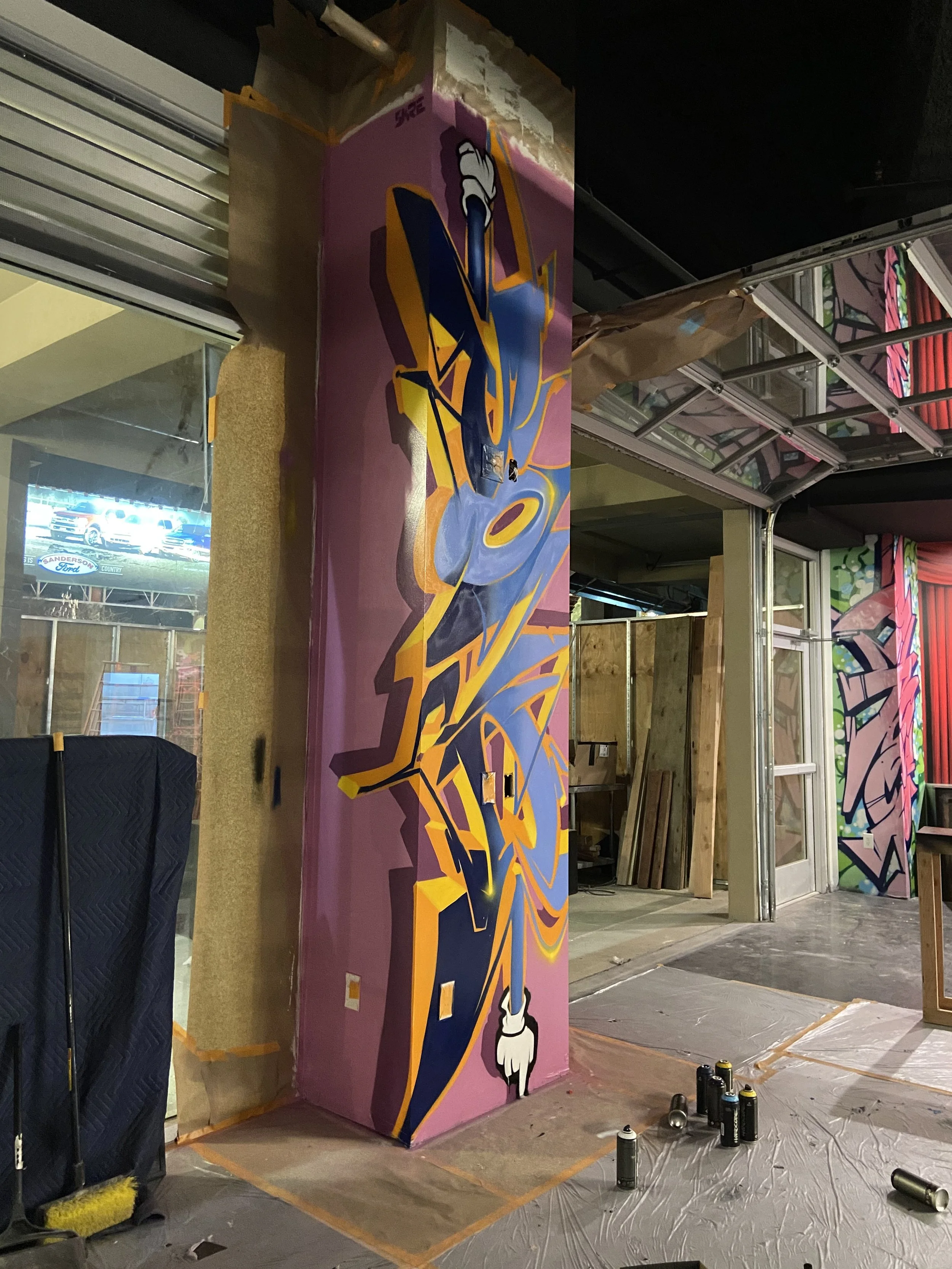 A colorful graffiti mural being painted on a wall inside a building, with spray paint cans on the floor, and a partially covered doorway on the right.