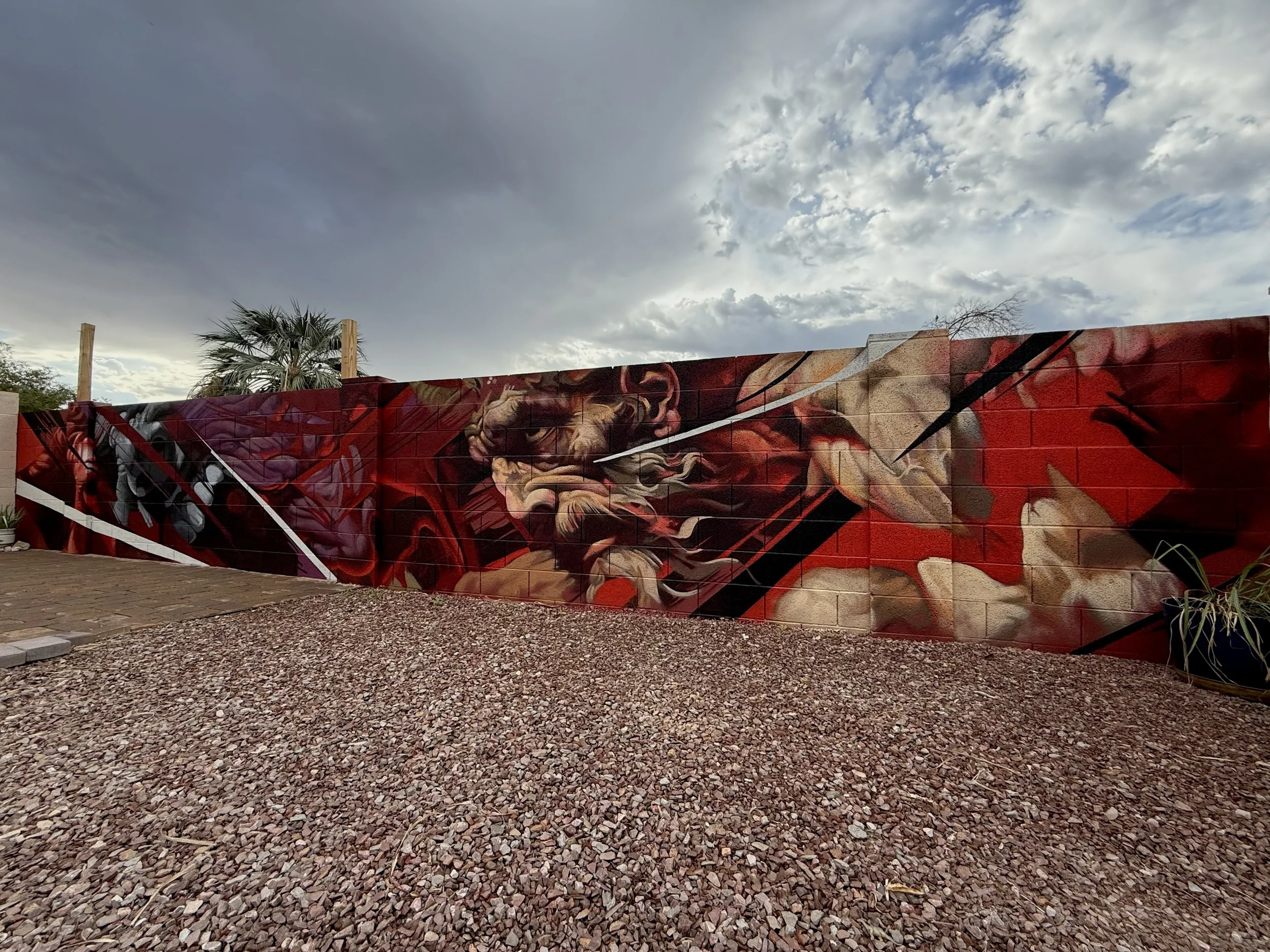 A mural painted on a red brick wall featuring a detailed close-up of a man transforming into Dracula face with a dark background and abstract lines and screaming faces, with a cloudy sky above and a tree in the distance.