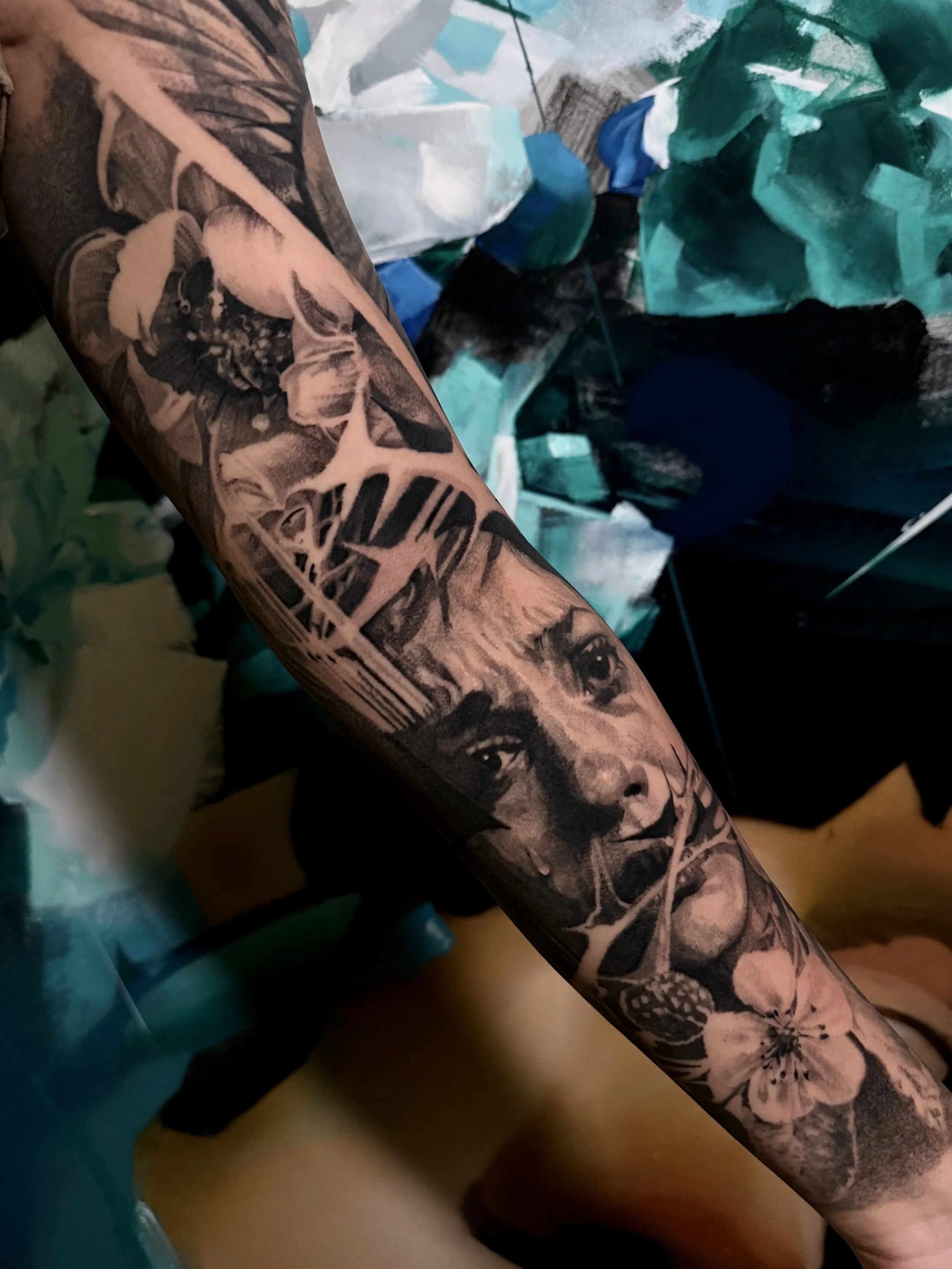 A tattooed arm with detailed black and gray tattoos of faces, flowers, and thorns, with a background of colorful objects and wrappers.