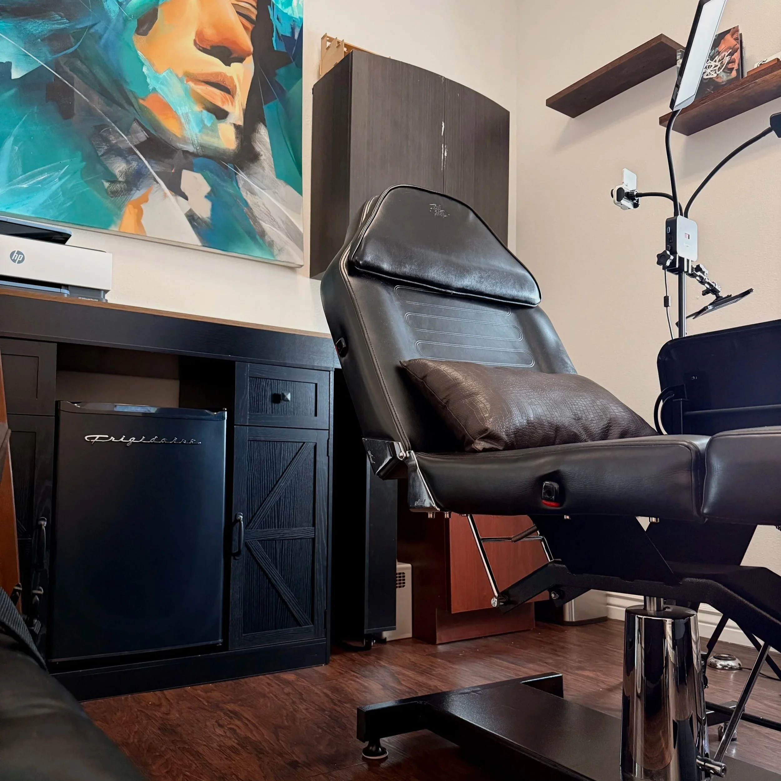 A tattoo studio setup with a colorful abstract portrait painting on the wall, a black leather tattoo chair, a wooden desk with a small refrigerator, cabinet, a TV mounted on a wooden shelf, and a light-colored wall and hardwood floor.