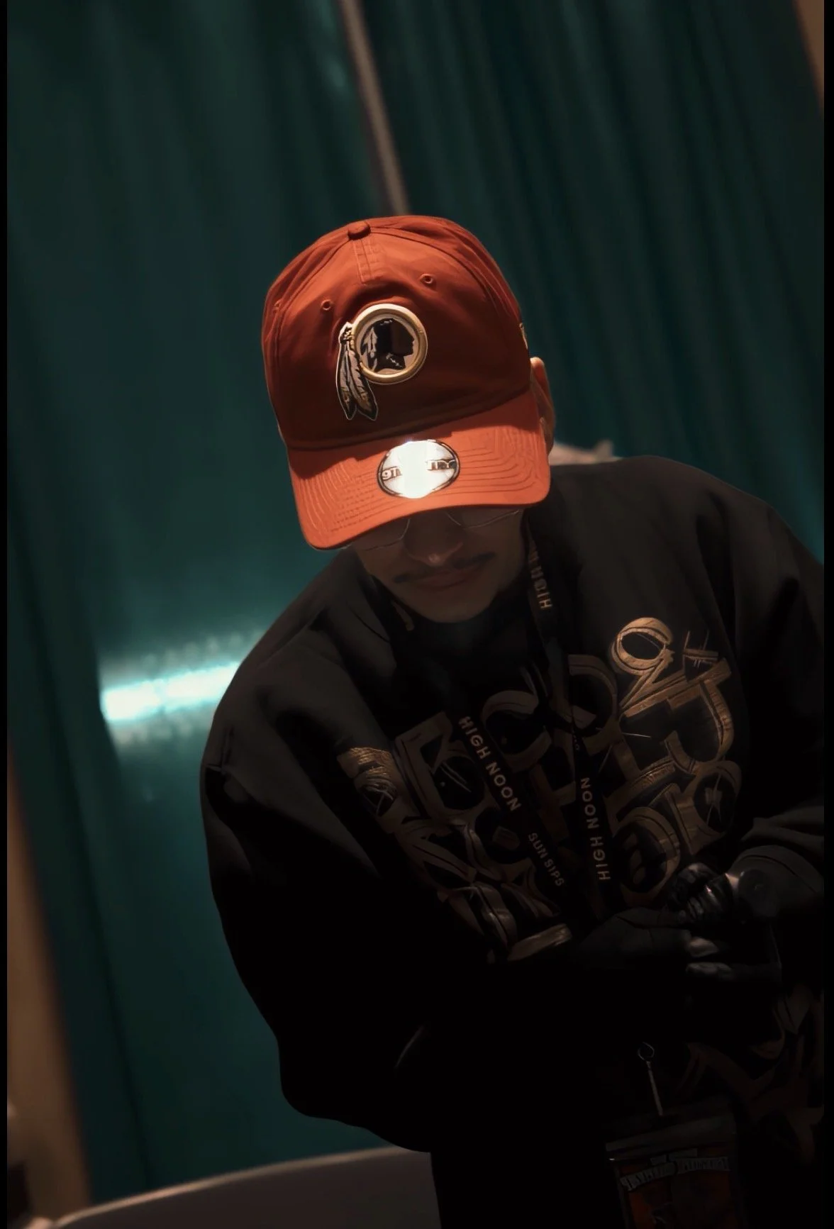 A person wearing an orange baseball cap with a military emblem patch, a black jacket with various designs and text, and black gloves. The person appears to be focused, with downward gaze, against a dark teal background.