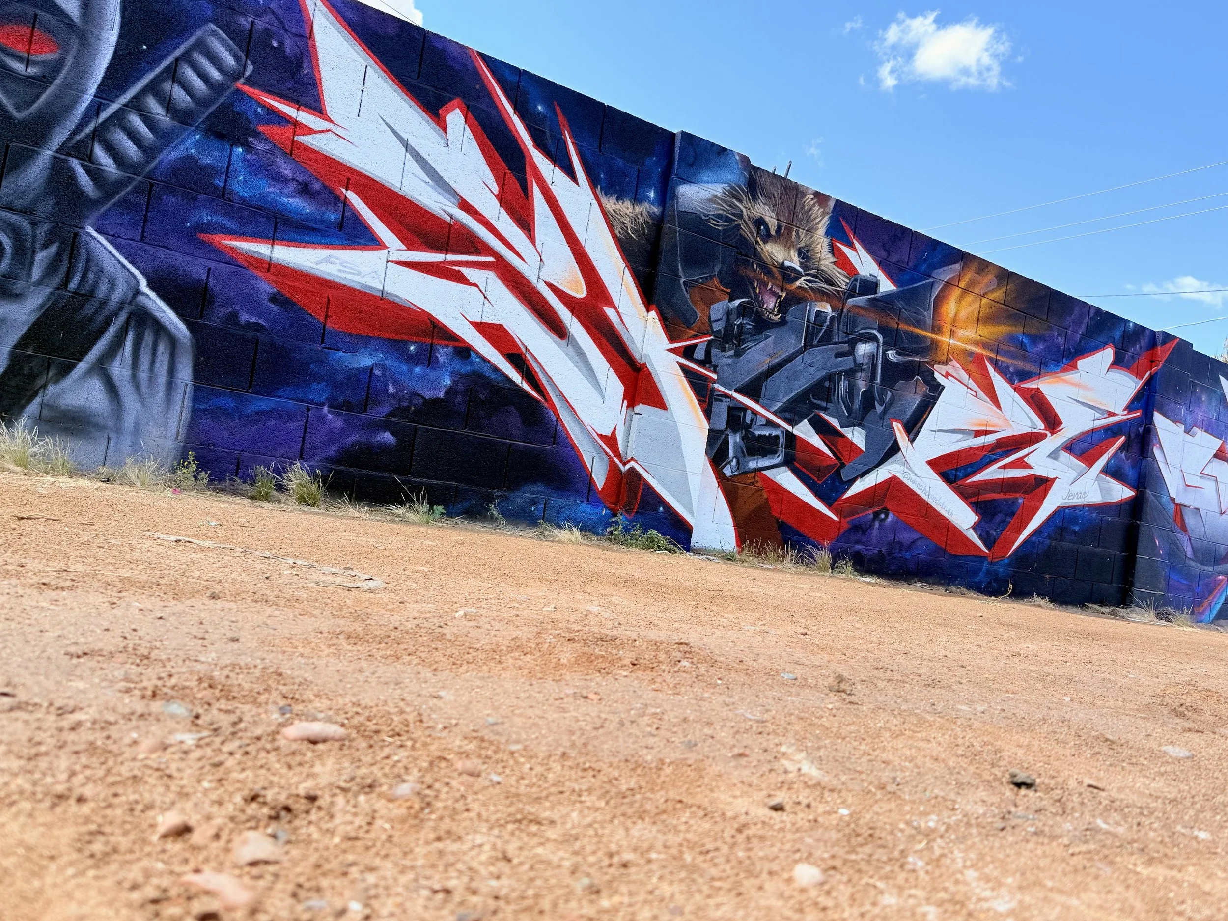 Colorful graffiti mural on a brick wall featuring Rocket from Guardians of the Galaxy, with nebula effects, and vibrant colors, with a blue sky background.