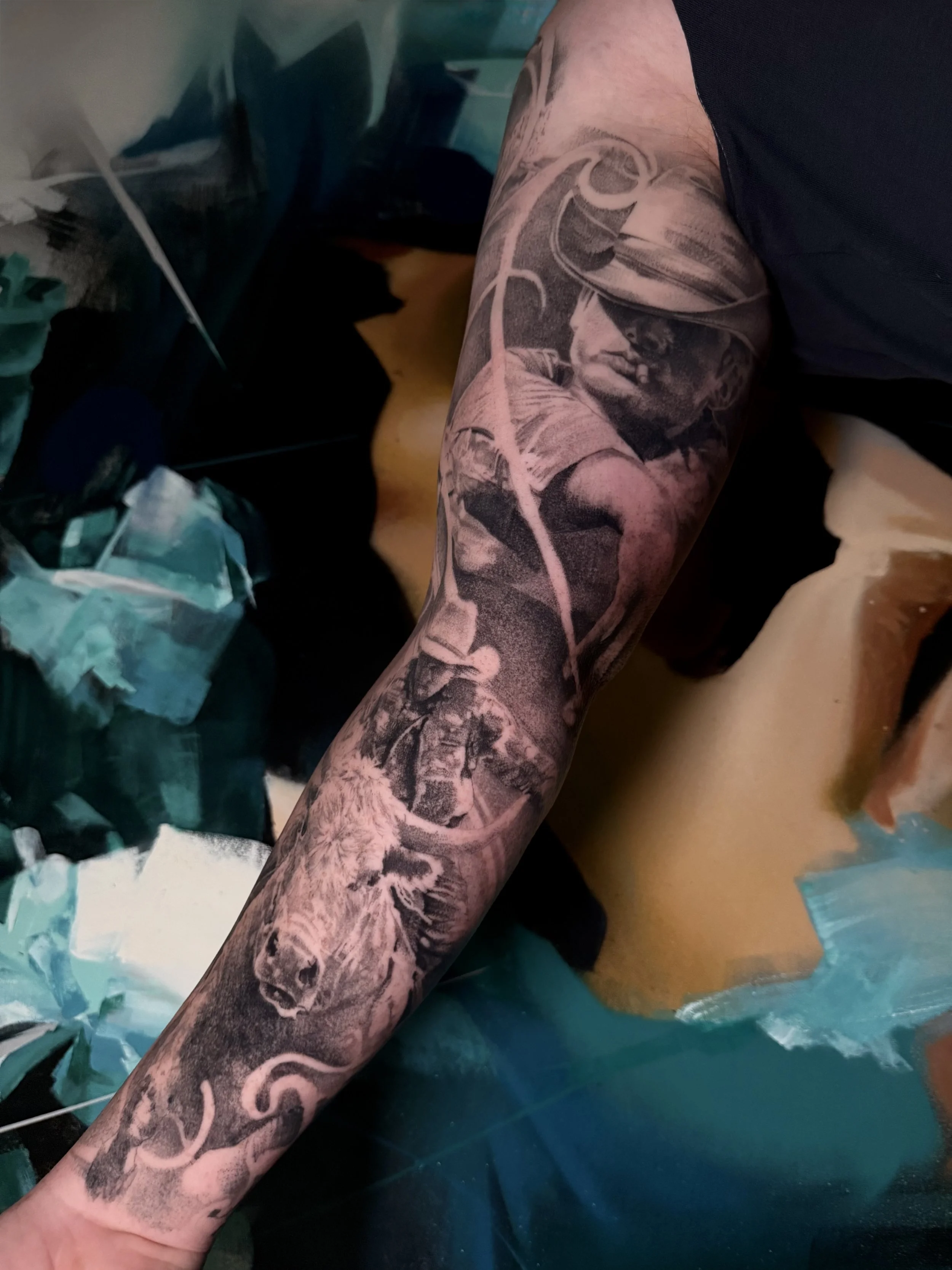 Close-up of a person's arm with a detailed black and gray tattoo featuring a cowboy wearing a hat, along with a bull rider, and skin break designs surrounding the image.