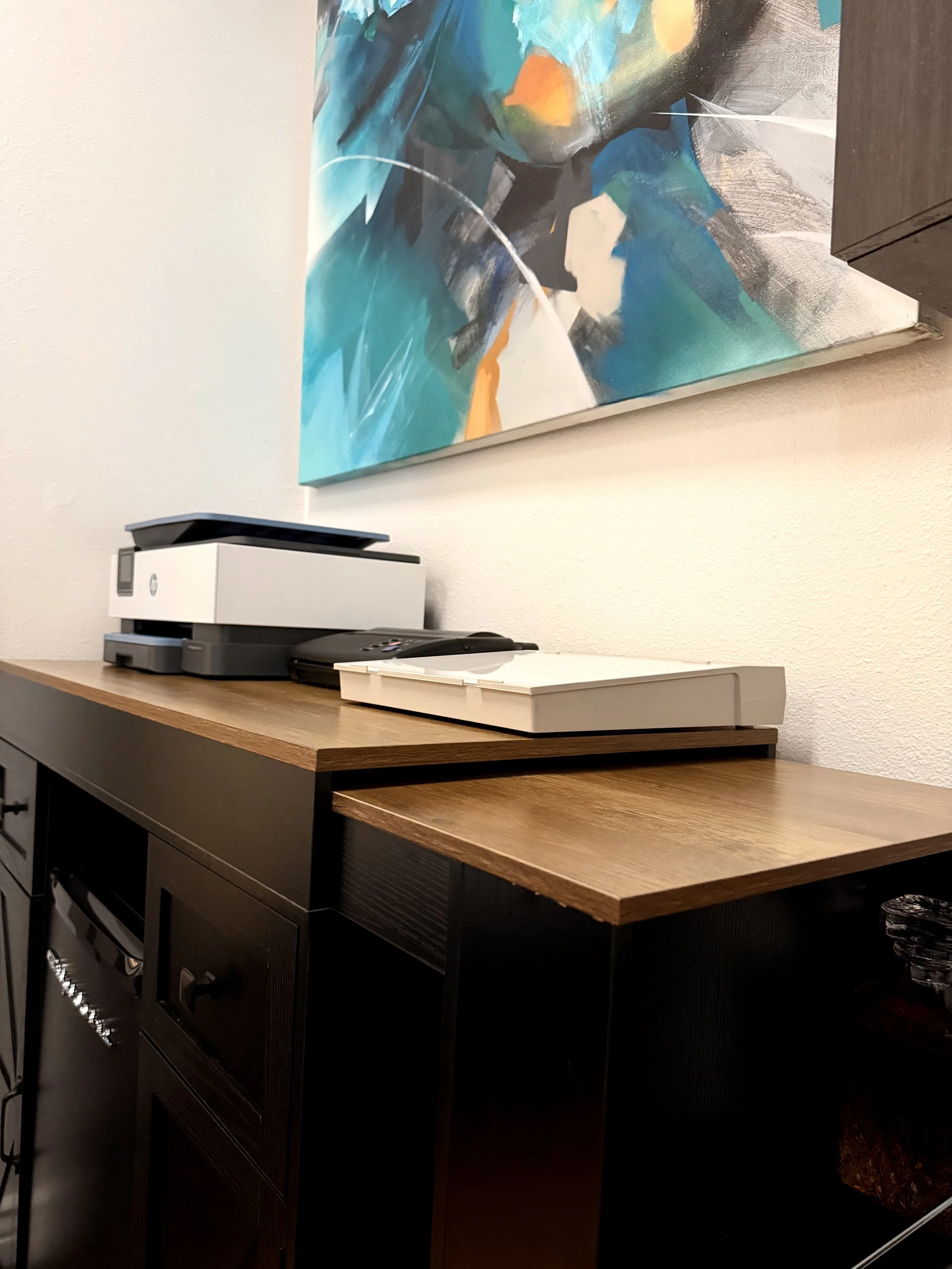 A wooden cabinet with a white printer on top, a black device below it, and a colorful abstract painting hanging on the wall behind in an office or workspace setting.