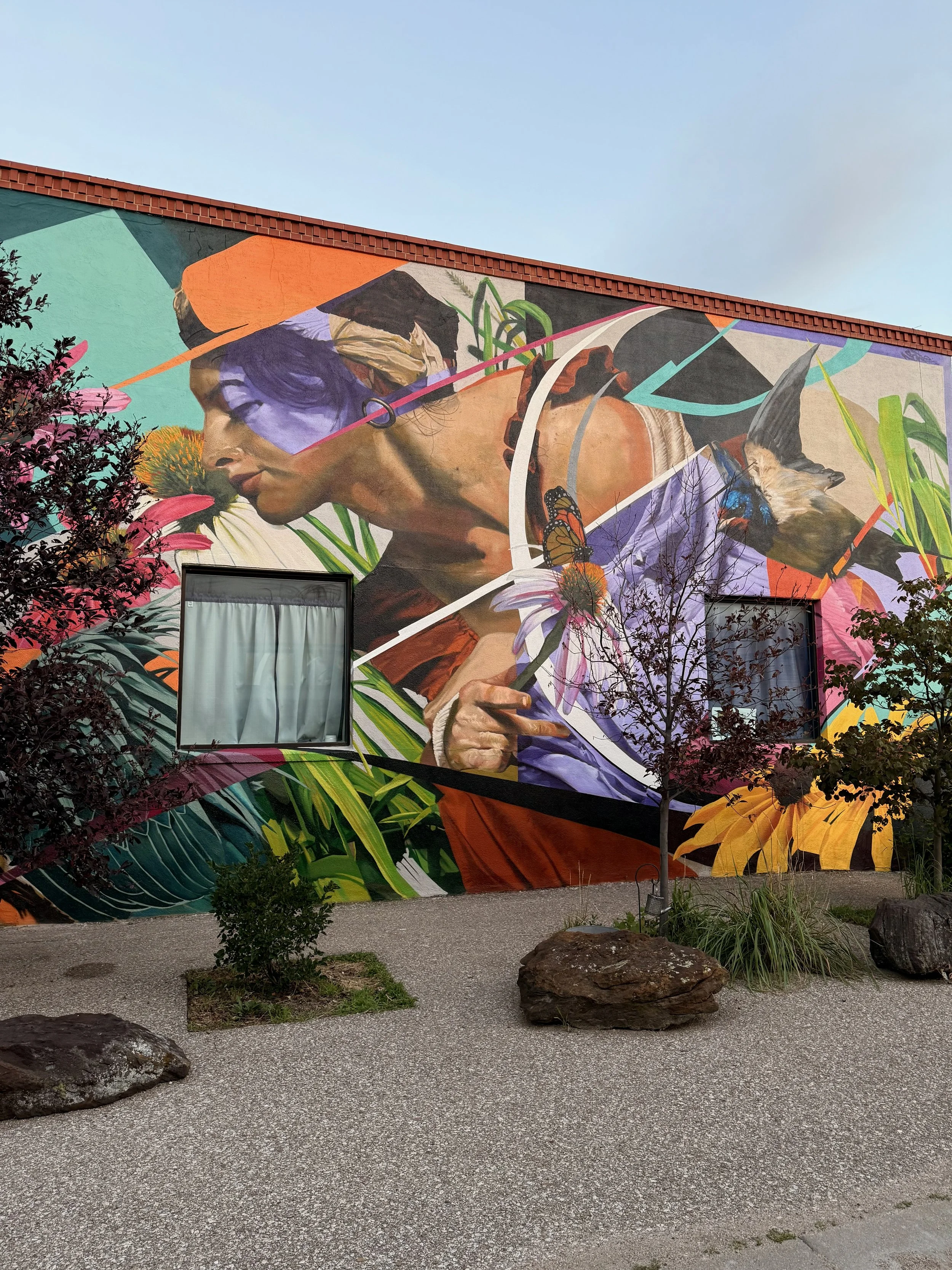 A large colorful mural on a building wall features a woman with short hair and a serene expression, surrounded by vibrant flowers, leaves, and butterflies, with a clear blue sky above.