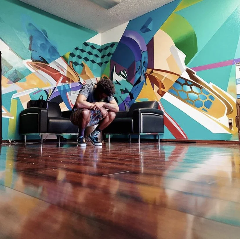 Young man sitting on a black leather sofa with a colorful abstract mural on the wall behind him, in a room with polished wooden floors.