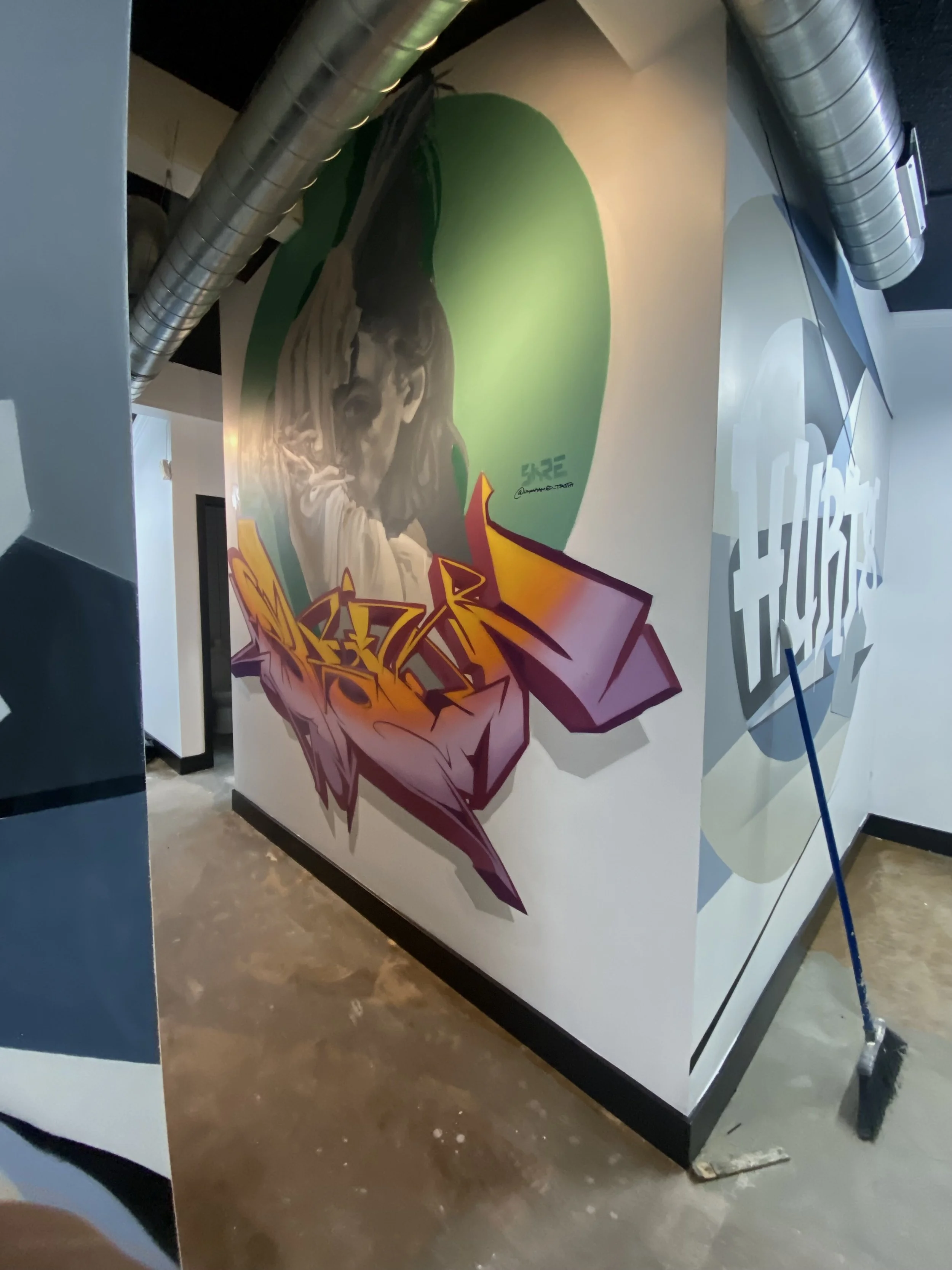 A wall mural featuring a stylized portrait of a woman holding up her hair, painted with vibrant colors and graffiti-style lettering underneath, located in an indoor space with exposed ductwork and a polished concrete floor.