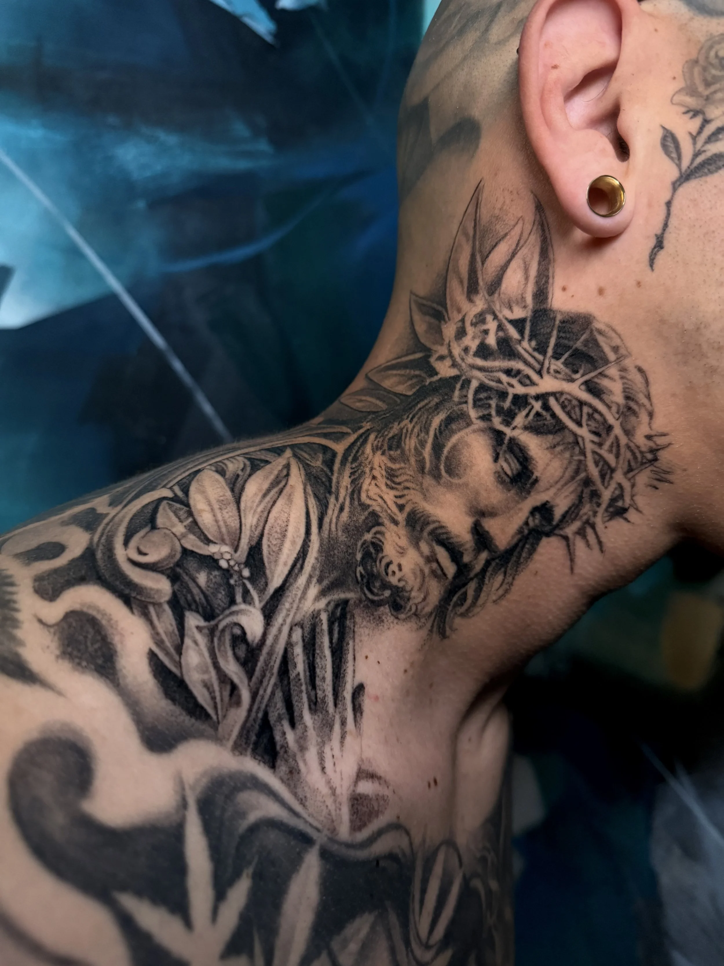 Close-up of a person's upper arm and neck showing extensive black ink tattoos, including a Jesus portrait, leaves, and abstract shapes.