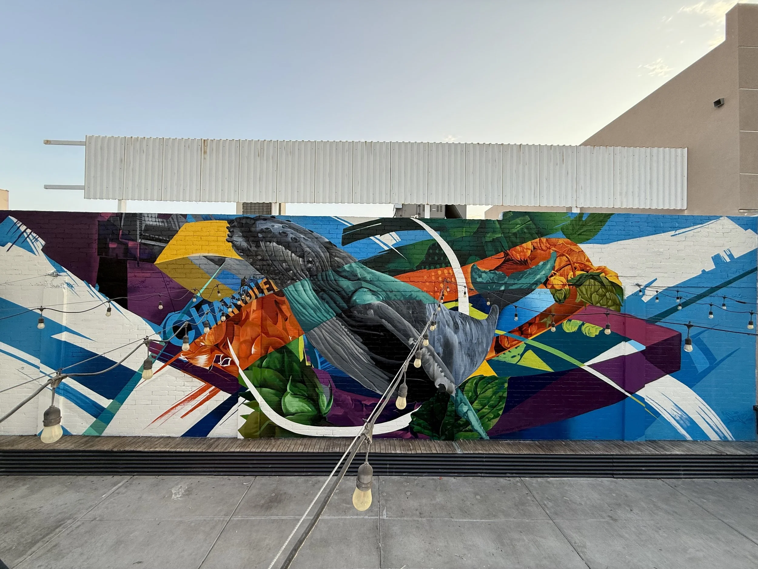 Colorful mural on a white brick wall featuring abstract shapes, a whale, and beer hops.