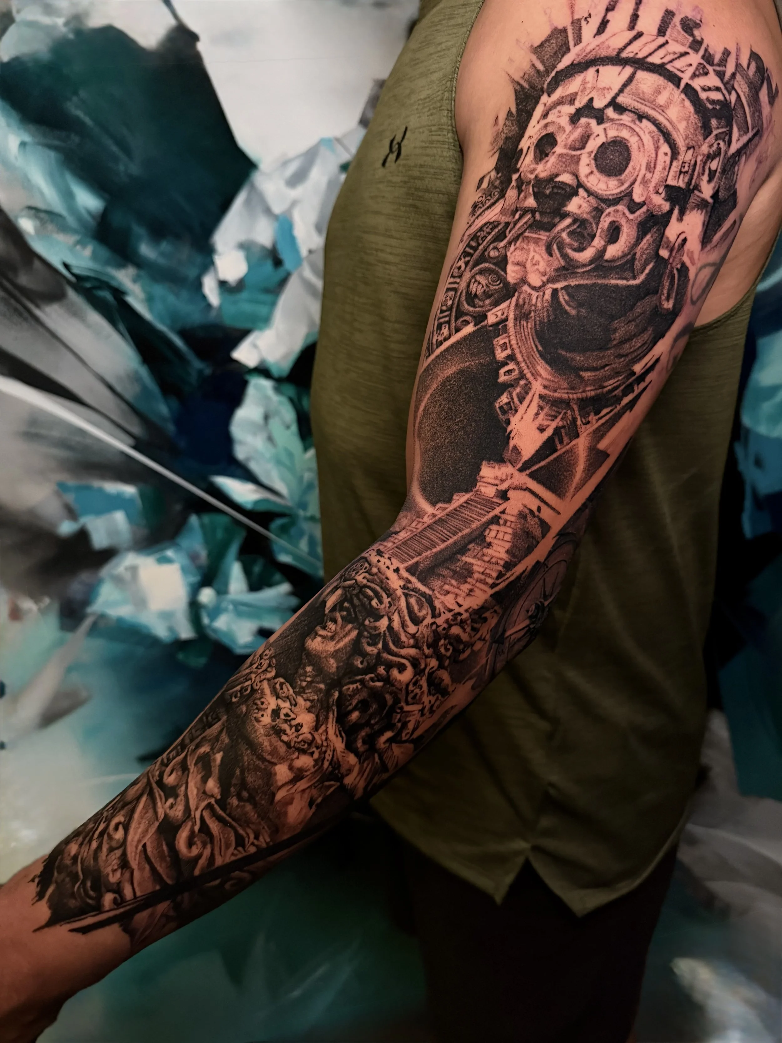 Close-up of a person's arm with a detailed black and gray tattoo sleeve, featuring Aztec elements with dynamic mark making.