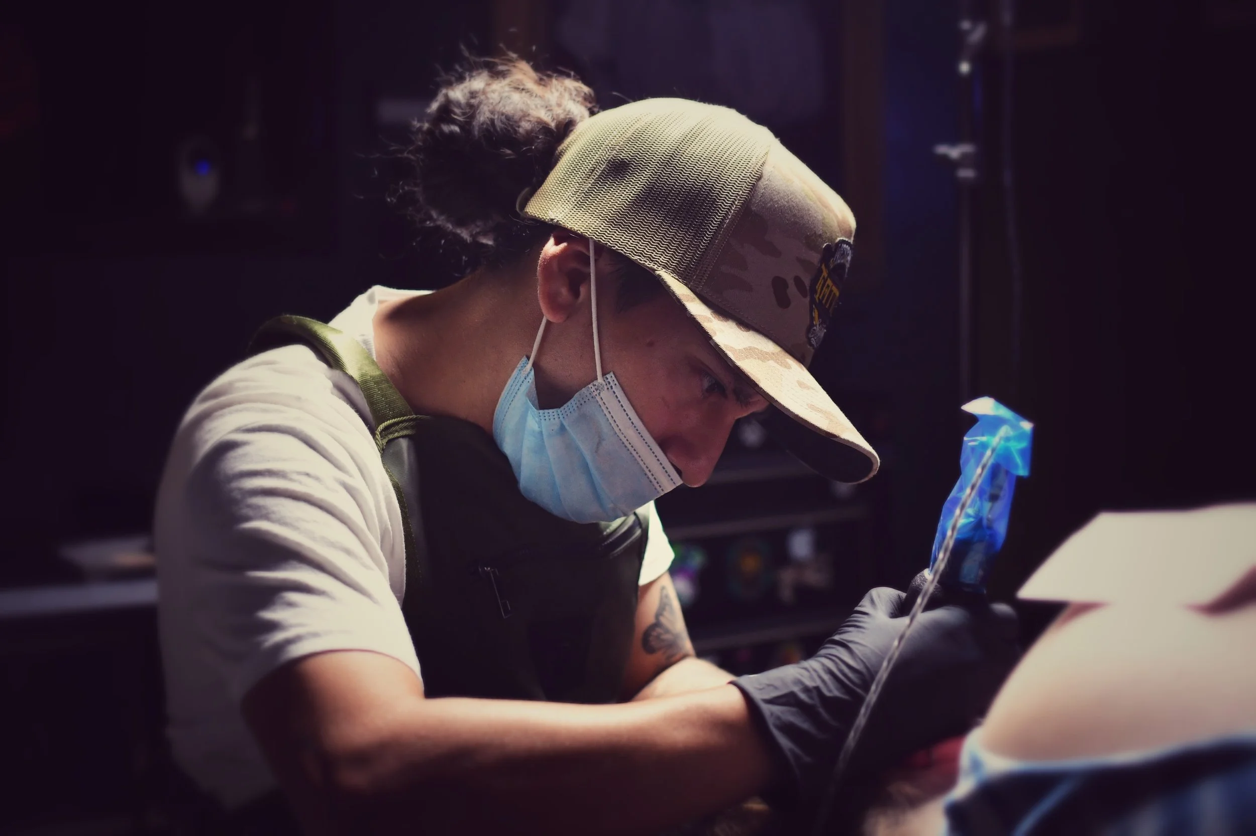 A person wearing a camouflage baseball cap and a face mask, focused on a task, with dark background and bright objects nearby.