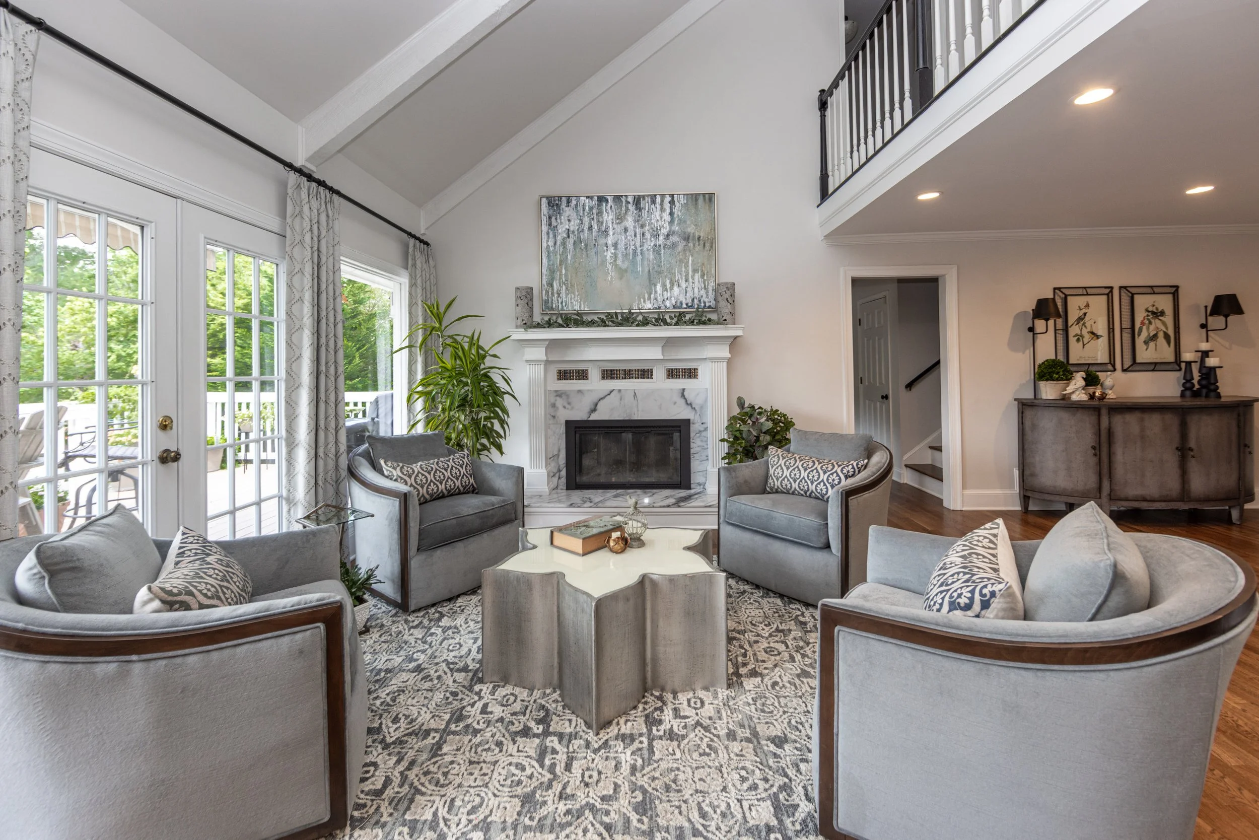 Living room with a white fireplace, abstract art above, multiple gray armchairs with patterned pillows, large windows with curtains, potted plants, and a cozy seating area.