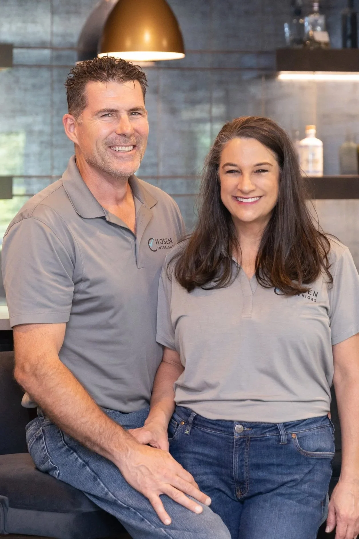 A smiling man and woman wearing matching gray shirts with a company logo, sitting inside a modern interior space with dark walls, pendant lighting, and shelves with decorative items.