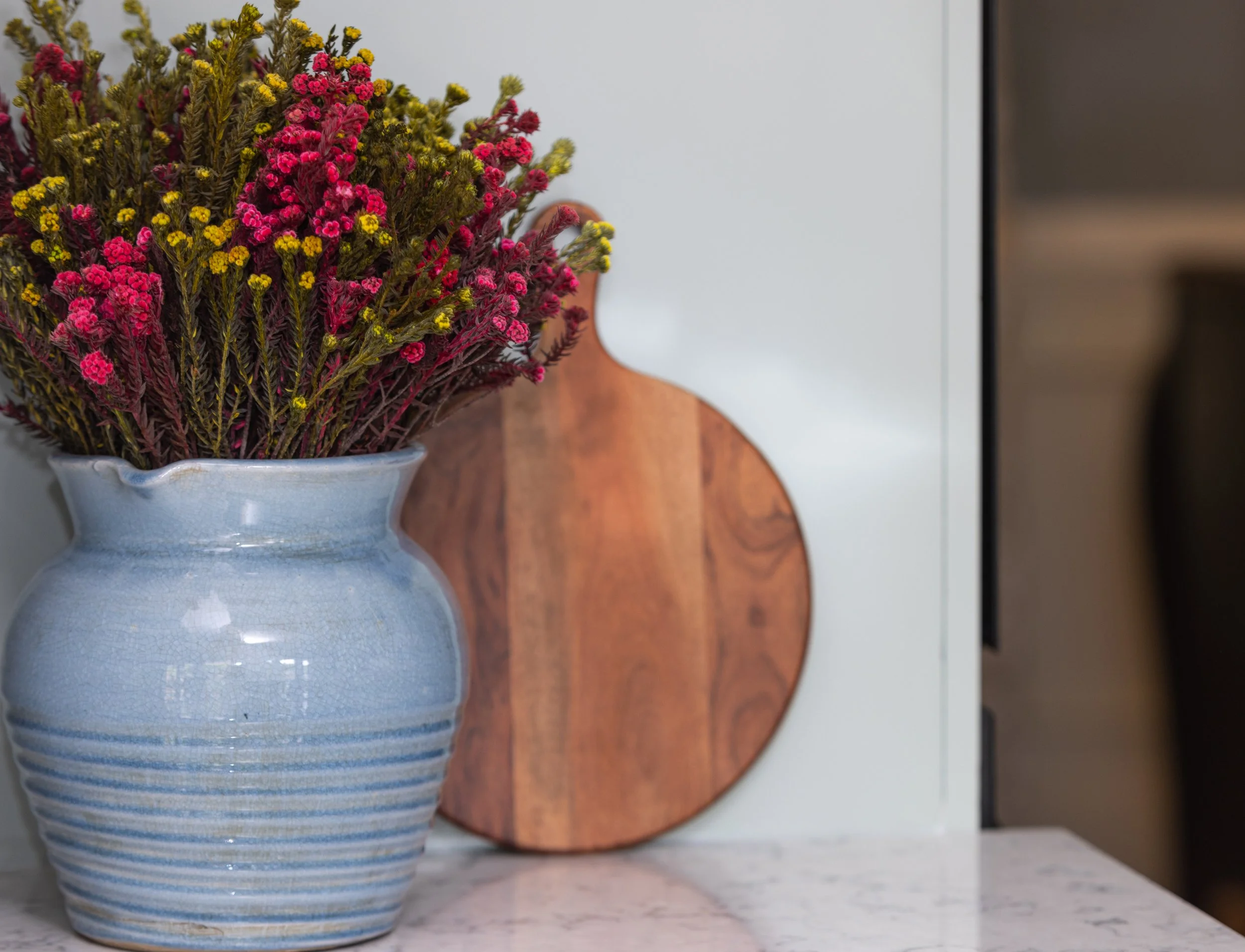 A white ceramic vase with blue stripes, holding a bouquet of red, pink, and yellow flowers. The vase is on a marble surface with a wooden object and a black-and-white background nearby.