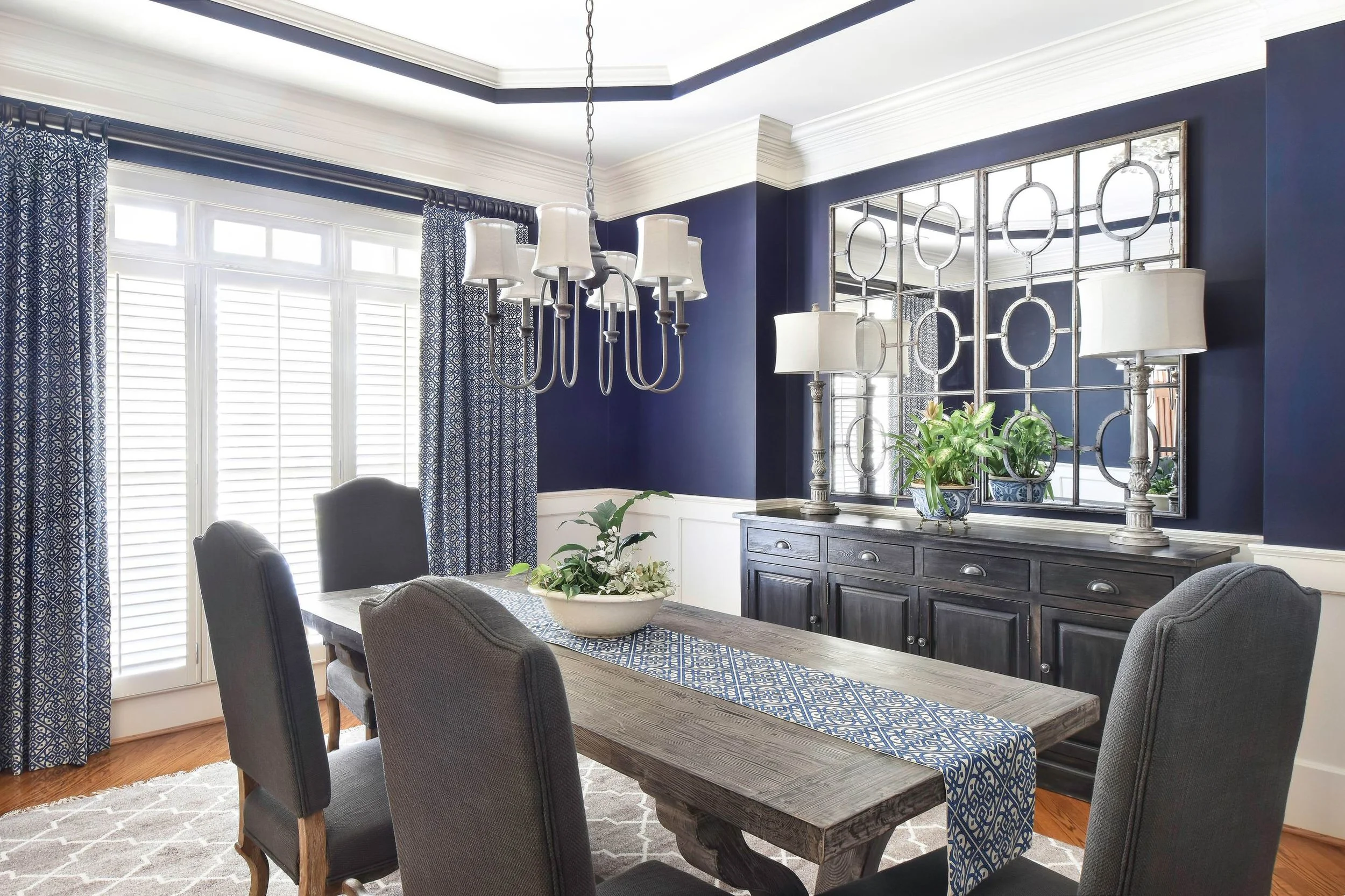 A dining room with navy blue walls, large windows with white plantation shutters, patterned blue and white curtains, a wooden dining table with a blue patterned runner, and six gray upholstered chairs. There is a black sideboard with silver hardware,