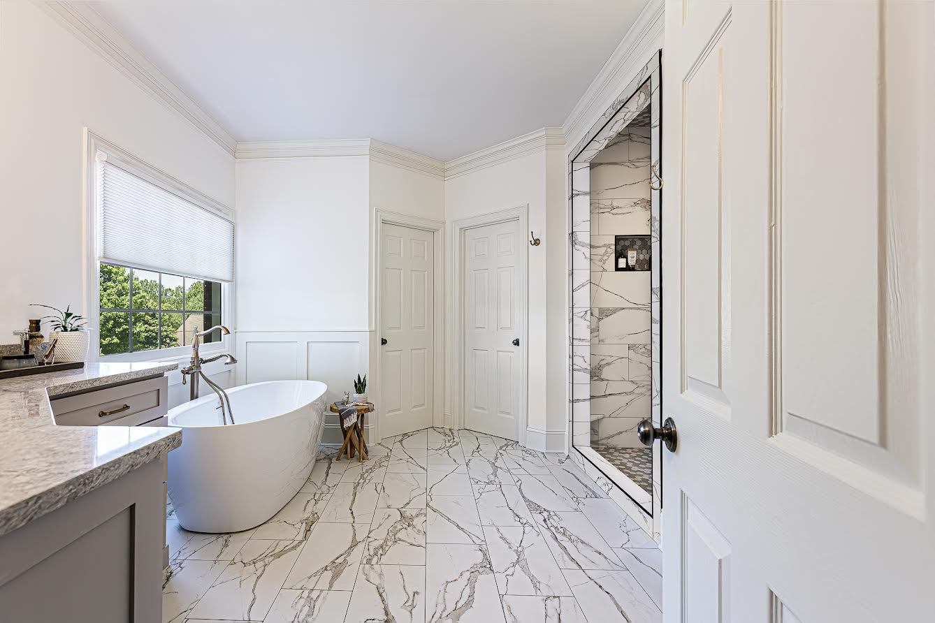 Bright bathroom with white walls, a freestanding bathtub, a marble tile shower, and marble floor tiles, with a window showing green trees outside.