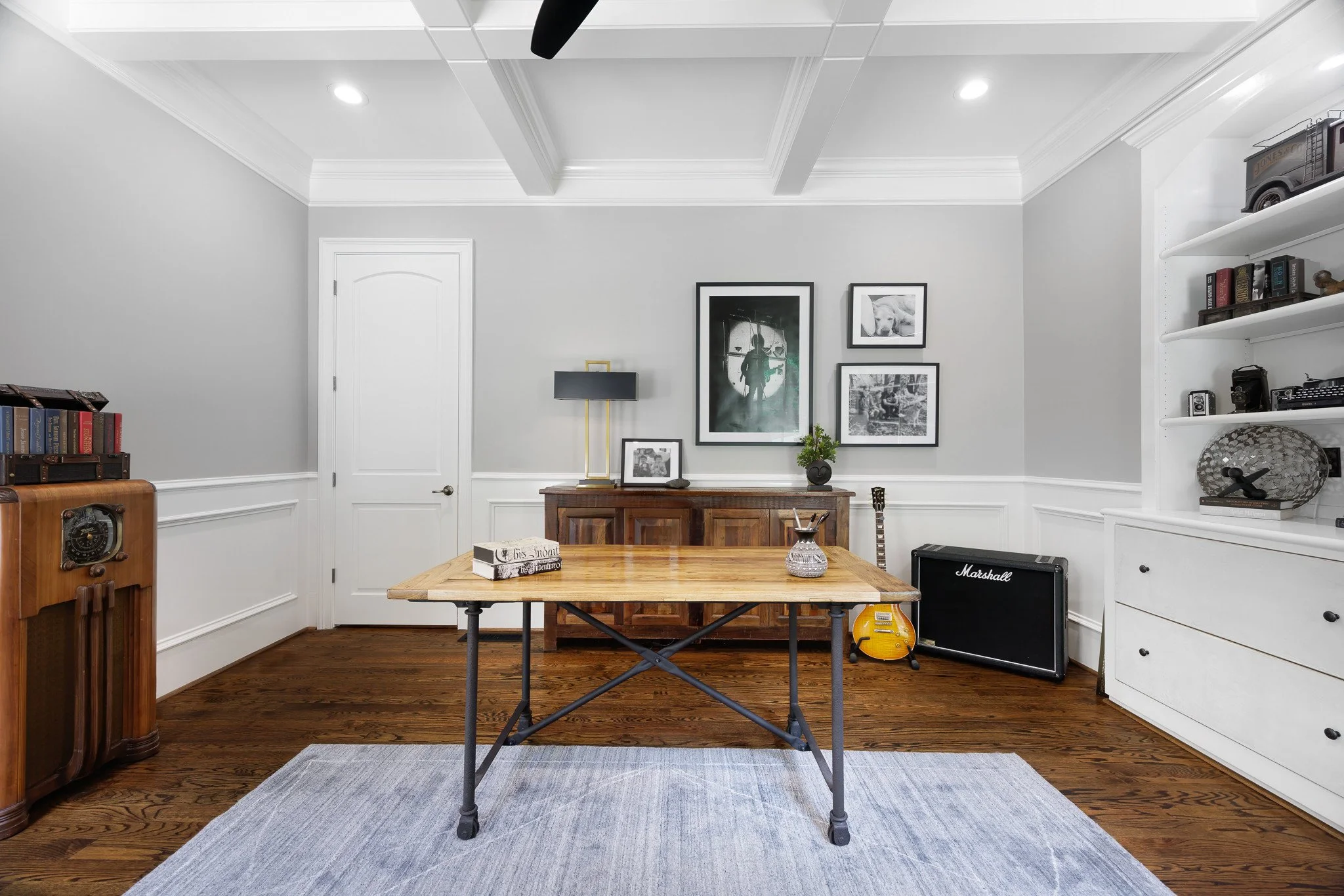 Interior of a room with gray walls and white trim, a wooden table with a black metal base, framed artwork on the wall, a white dresser, a guitar, and an amplifier.