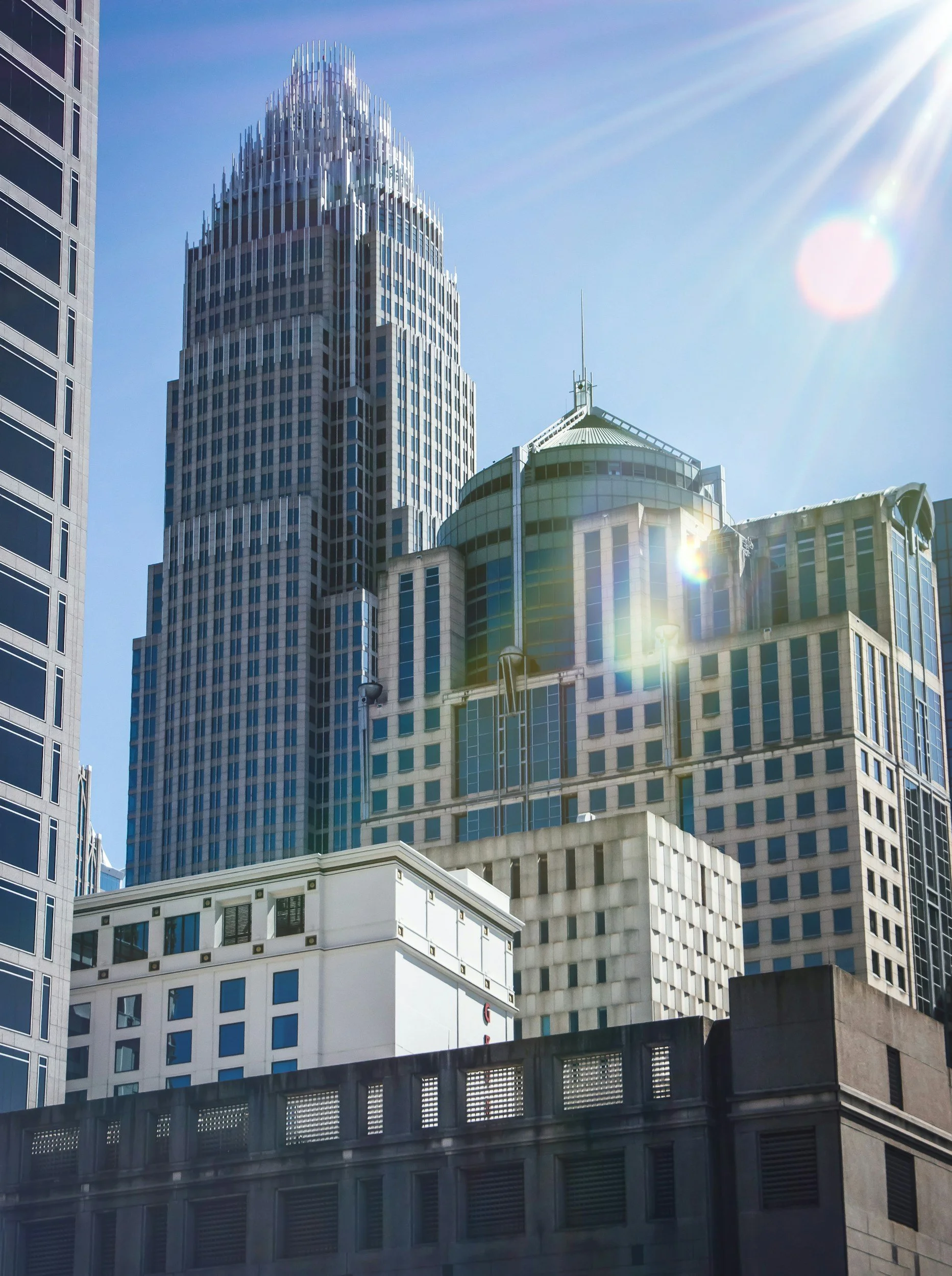 A cityscape with modern skyscrapers under a clear blue sky, with sunlight creating lens flare.