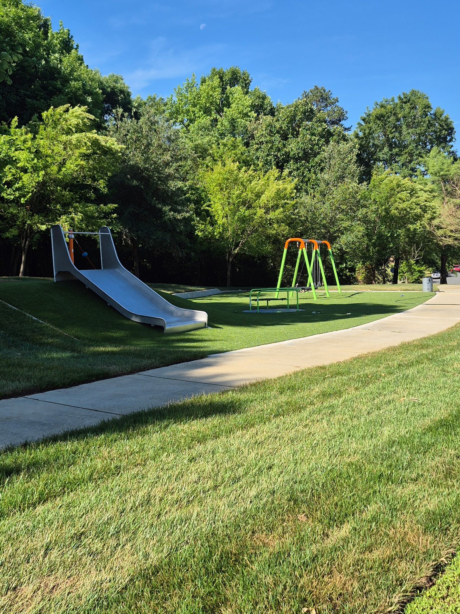 A playground with a slide, swings, and a bench on a grassy area, surrounded by tall trees and a clear blue sky.