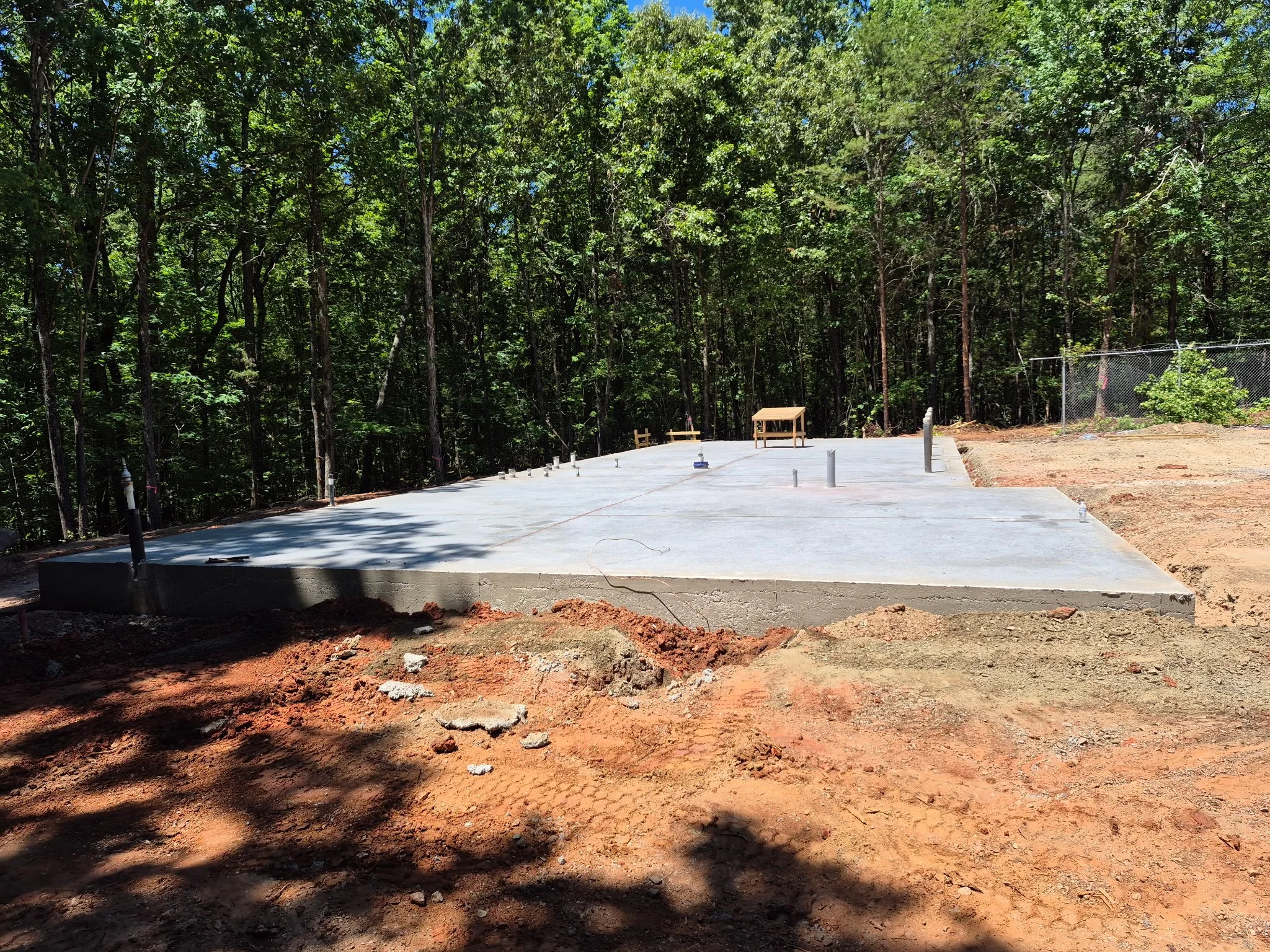Construction site with a concrete slab foundation, surrounded by dirt and soil, next to a chain-link fence, with a wooded area in the background.