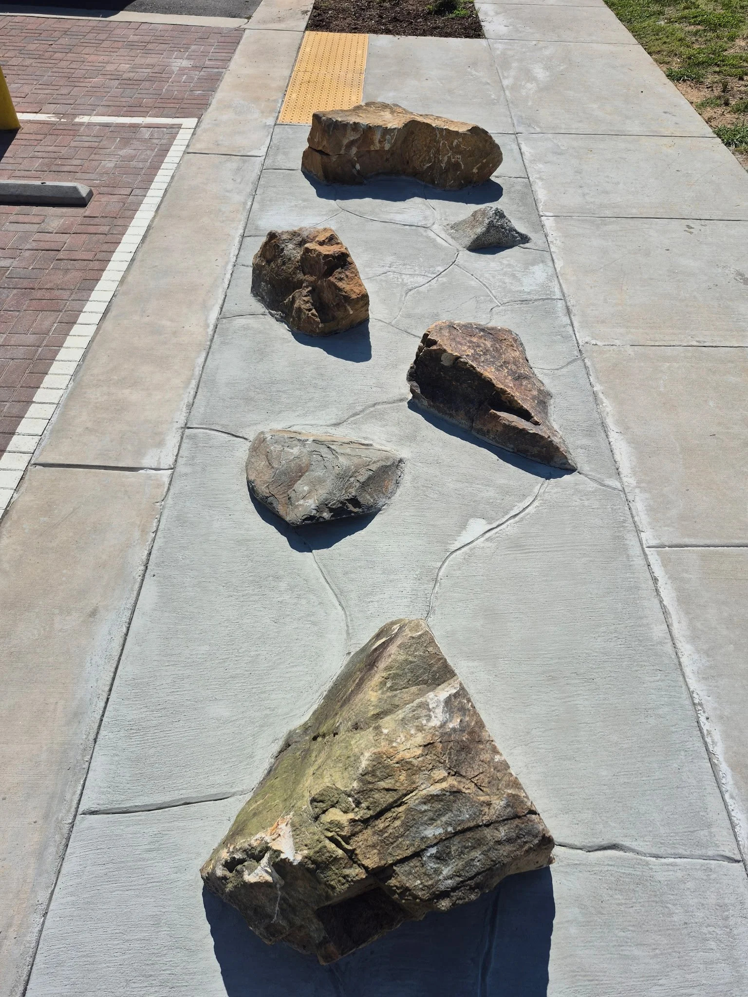Various large rocks arranged on a concrete sidewalk with a brick and asphalt surface nearby, and a patch of grass on the side.