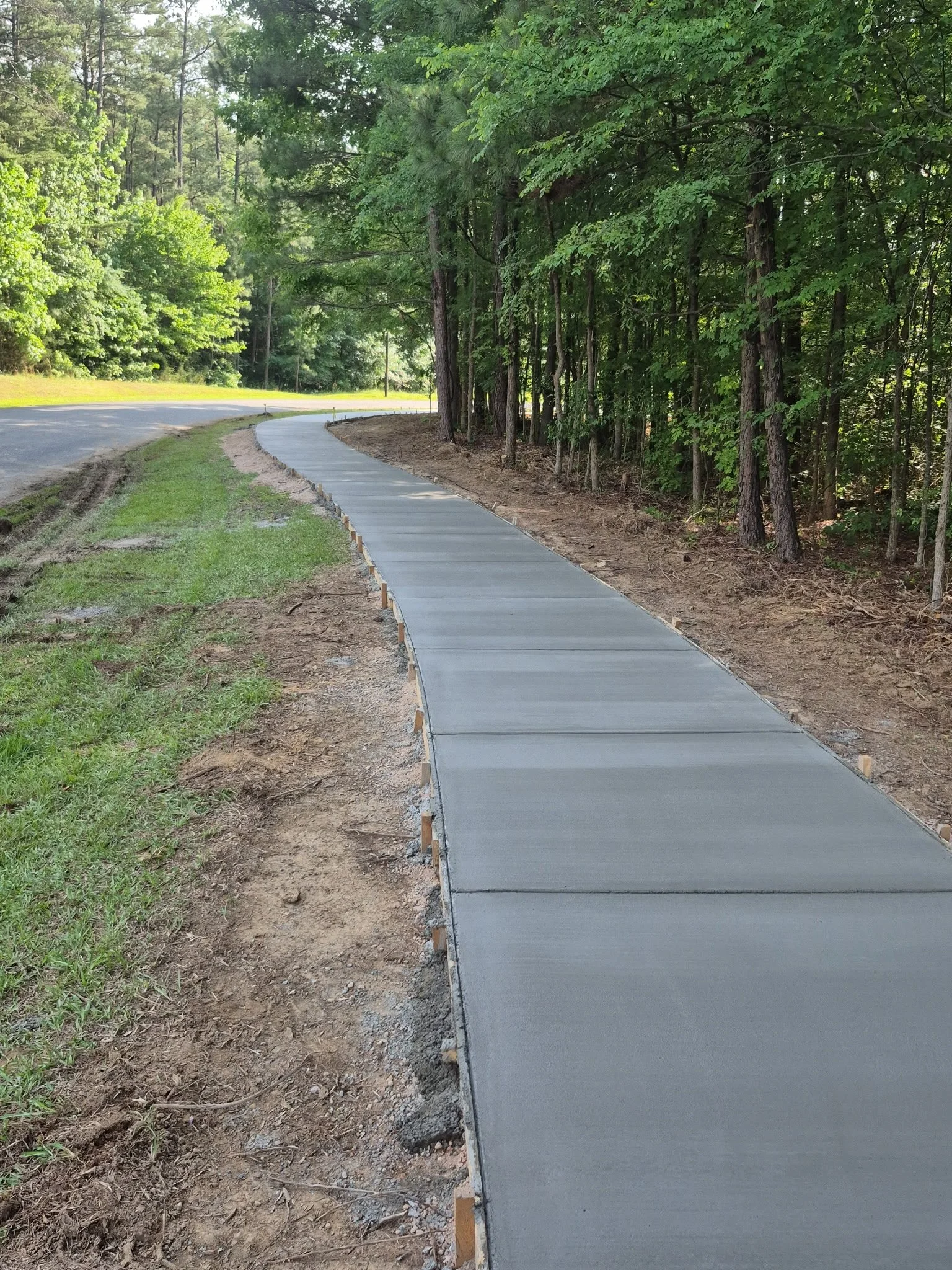 Newly poured sidewalk along a wooded area with trees on the right and grass on the left, with a paved road nearby.