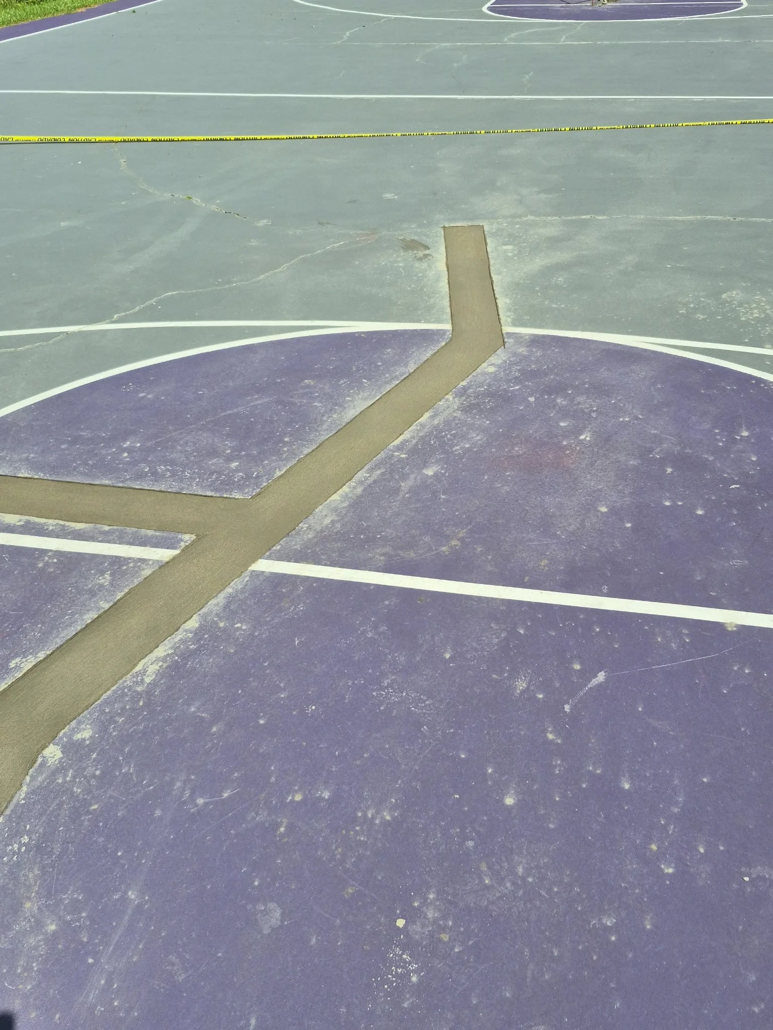 A worn basketball court with painted lines, cracked and scuffed surface, and a piece of cardboard or wood on the ground, with caution tape in the background.